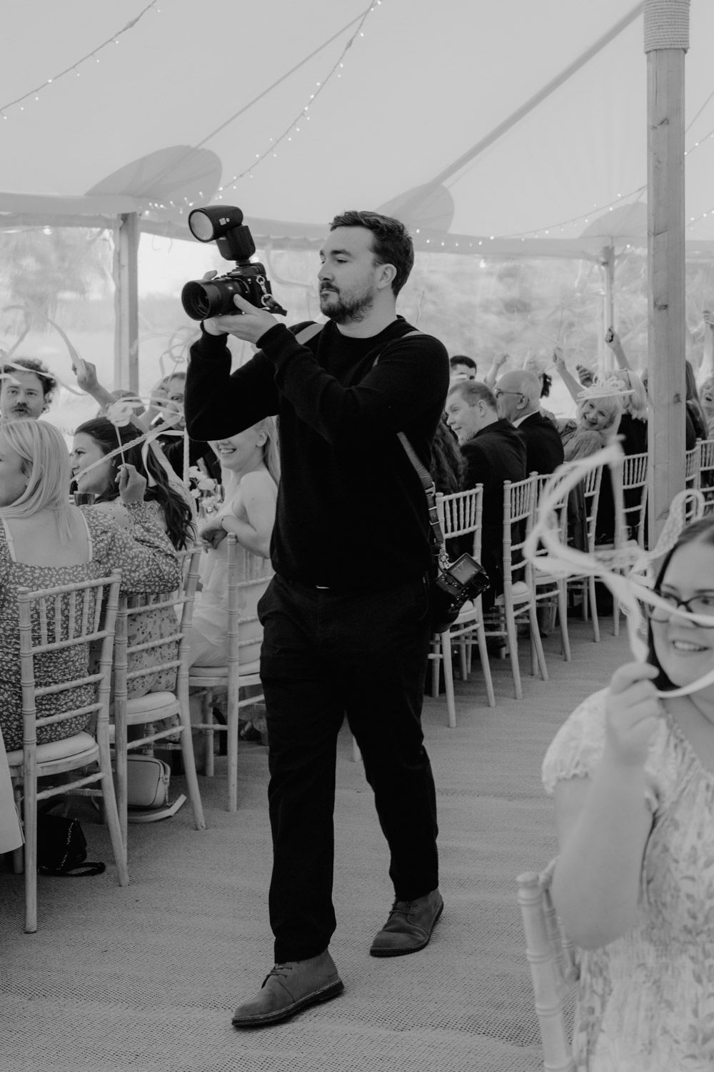 Black and white photo of a male photographer holding a camera at an elegant wedding reception in a marquee tent.