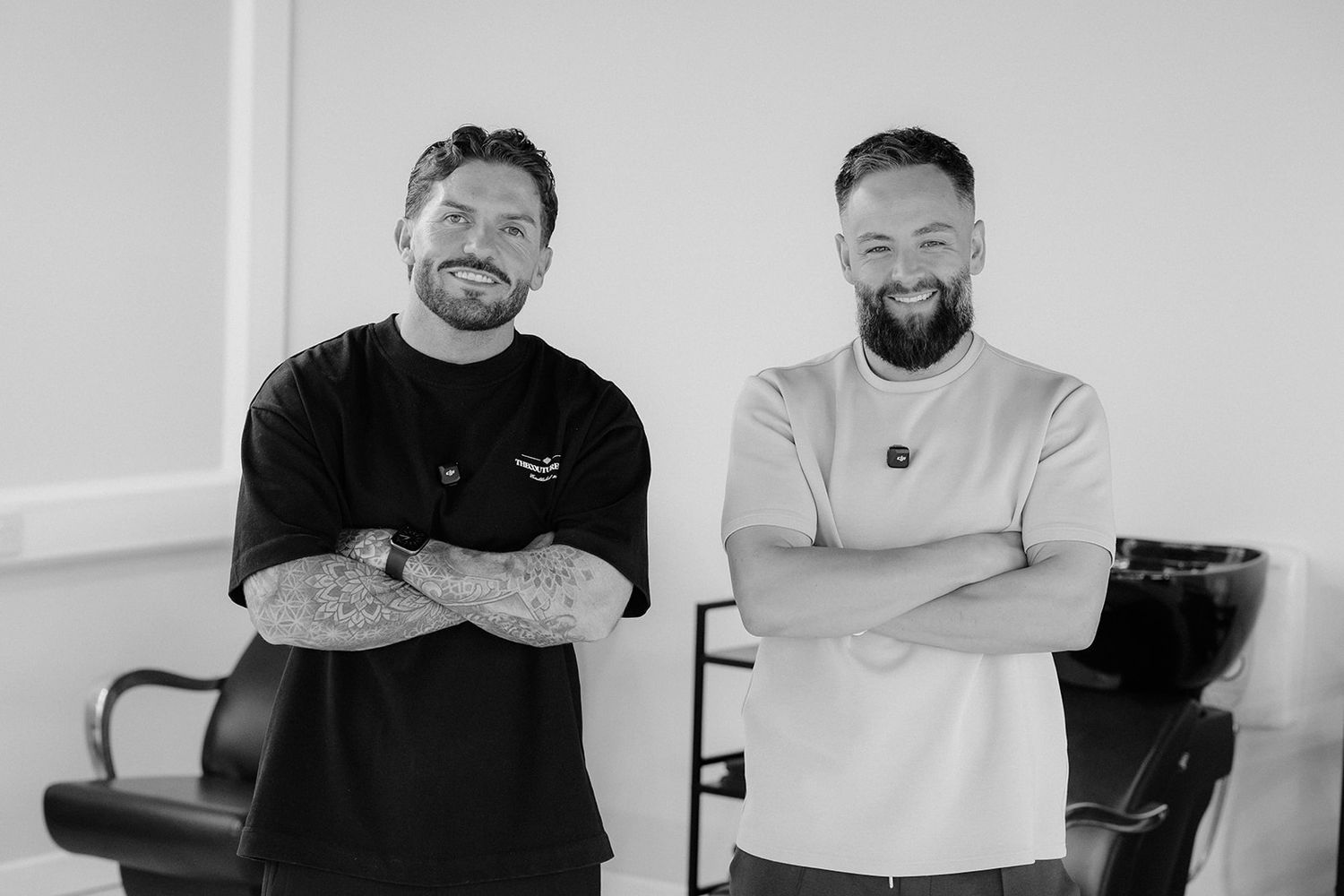 Two smiling men with beards and tattoos stand arms crossed in a modern barbershop, posing for a professional black and white photo.