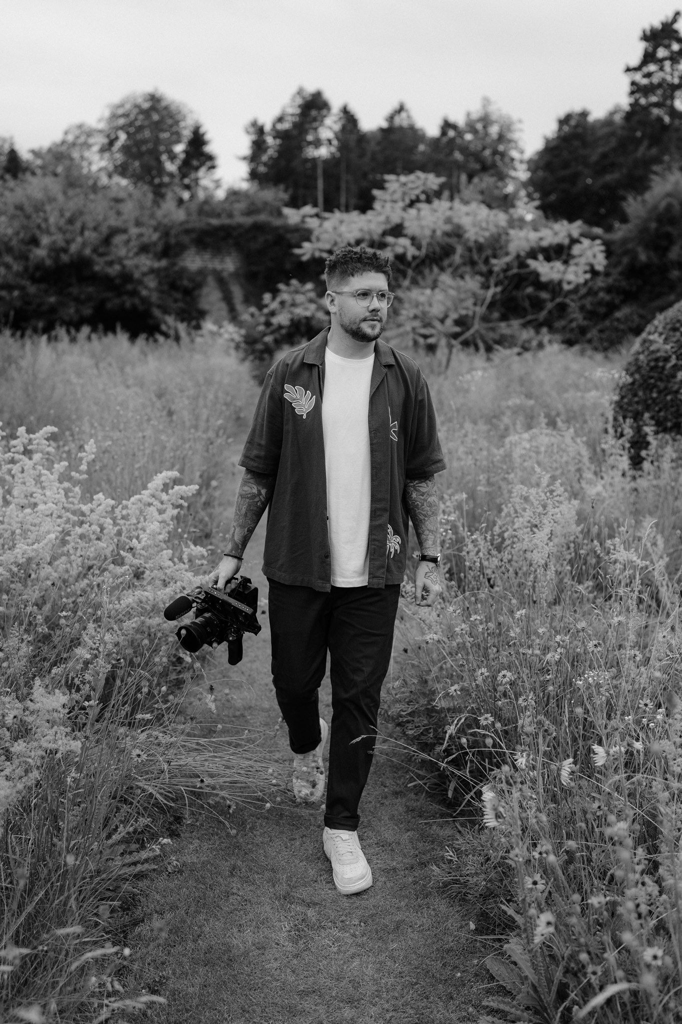 Black and white photo of a man walking on a nature path holding a camera, surrounded by wildflowers and trees.