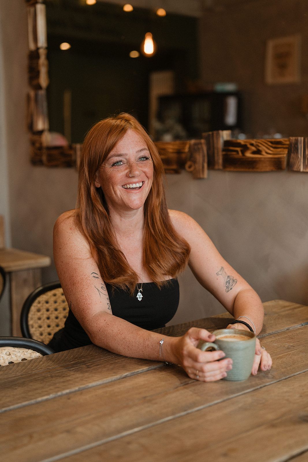 Smiling redhead woman with tattoos holding a latte mug at a wooden cafe table.