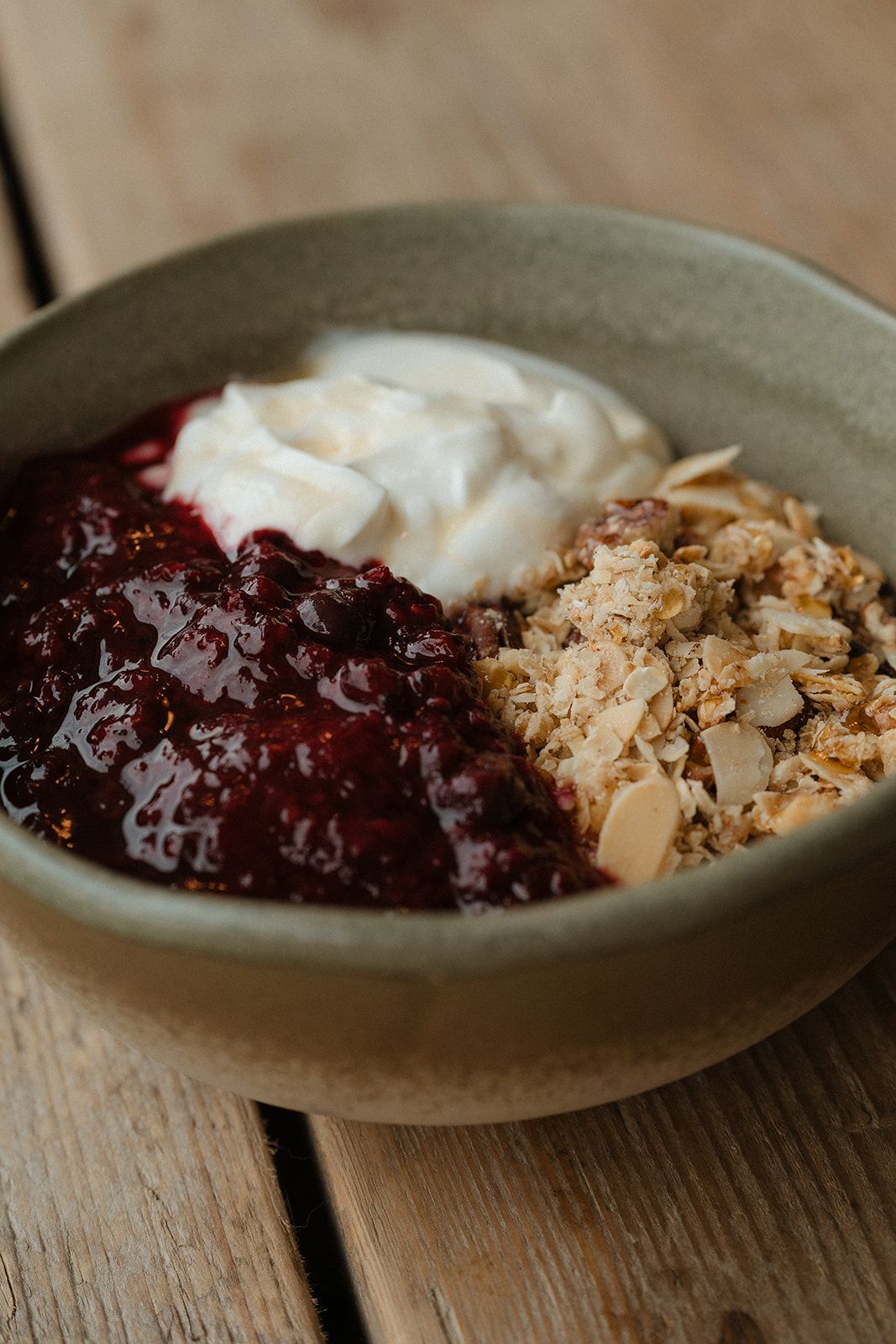 A ceramic bowl of granola with berry compote and creamy yogurt on a wooden surface.