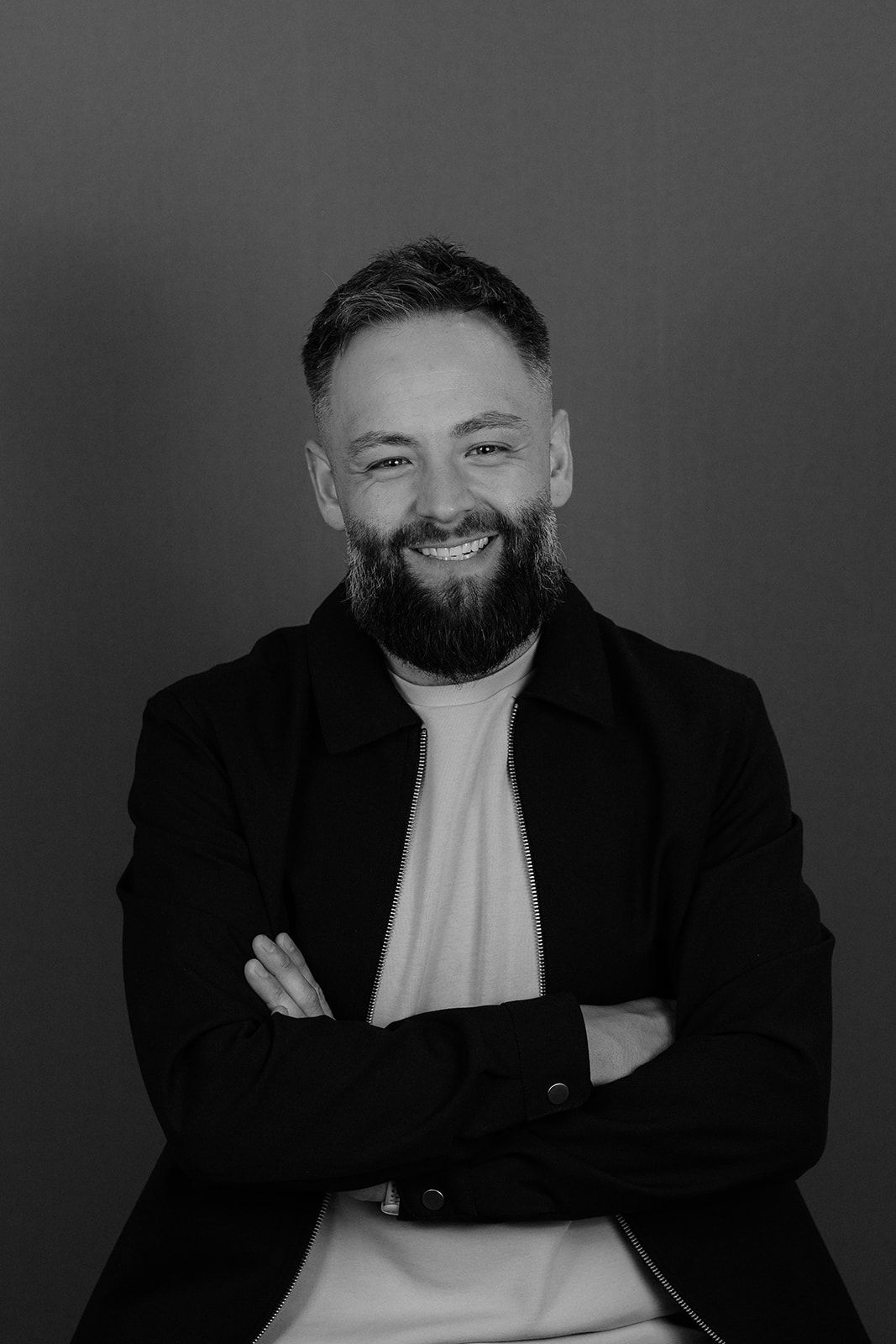 Smiling bearded man in black jacket with arms crossed poses confidently against gray background in black and white photo.