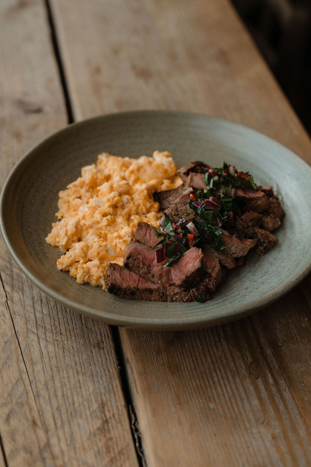 Sliced grilled steak with chimichurri sauce served alongside creamy scrambled eggs on a rustic ceramic plate.