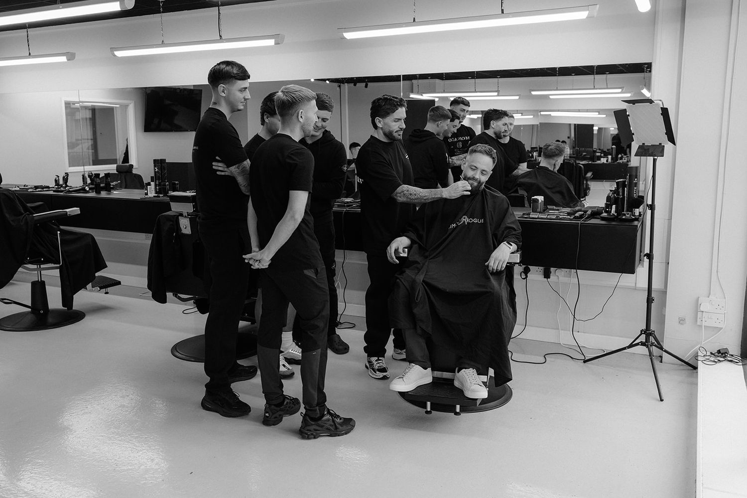 Black and white photo of barbers training in a barbershop, with one student cutting a clients hair.