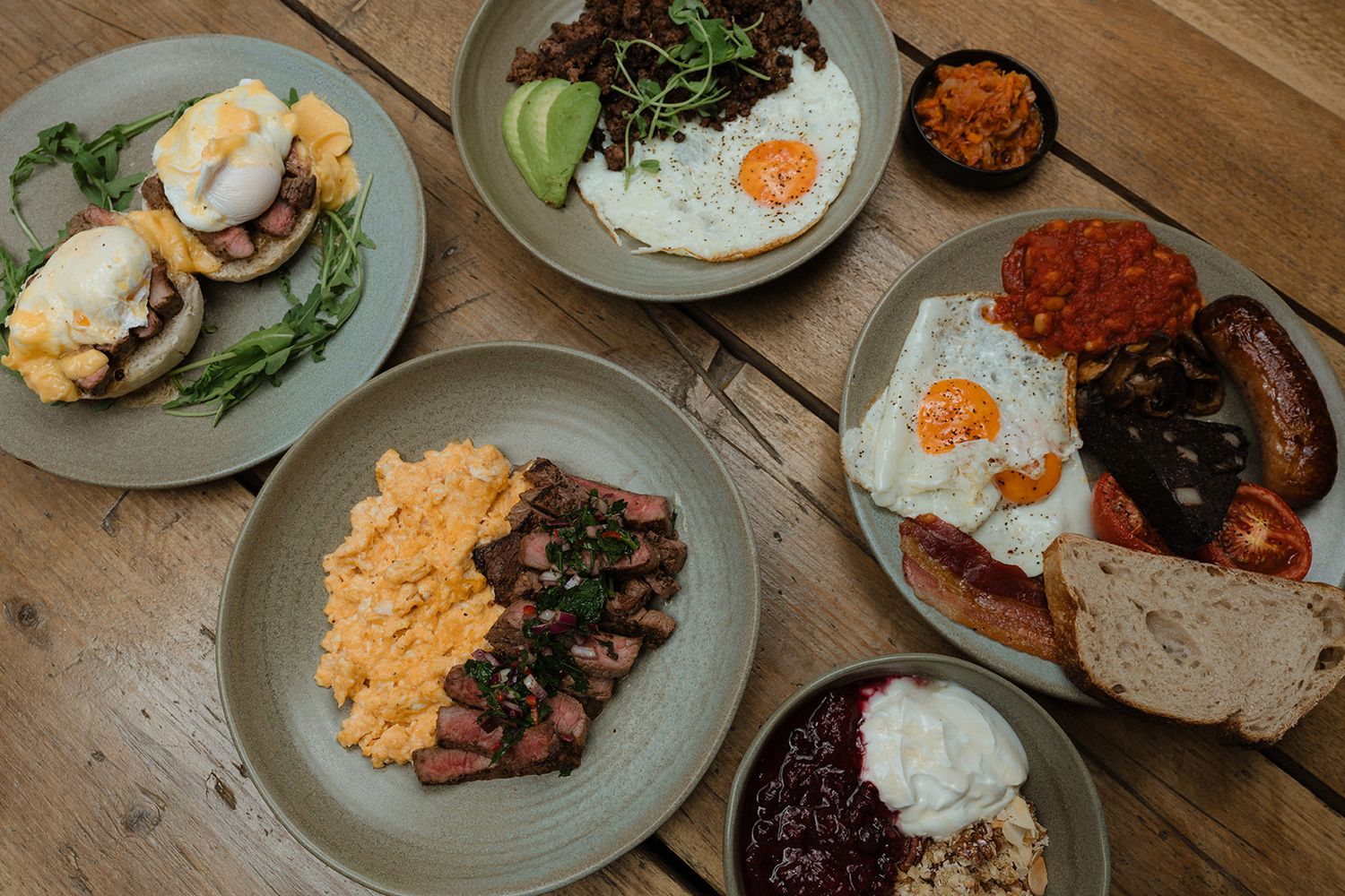 A spread of gourmet breakfast dishes including eggs benedict, steak with scrambled eggs, and a full English on a wooden table.