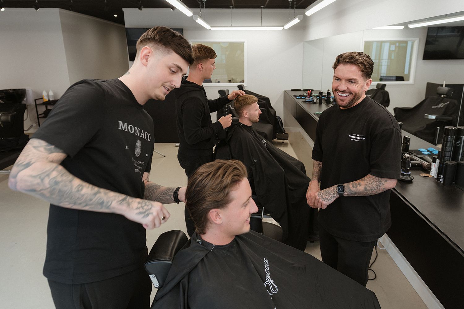 Two barbers in black uniforms cutting hair and smiling in a modern barbershop with white walls and mirrors.