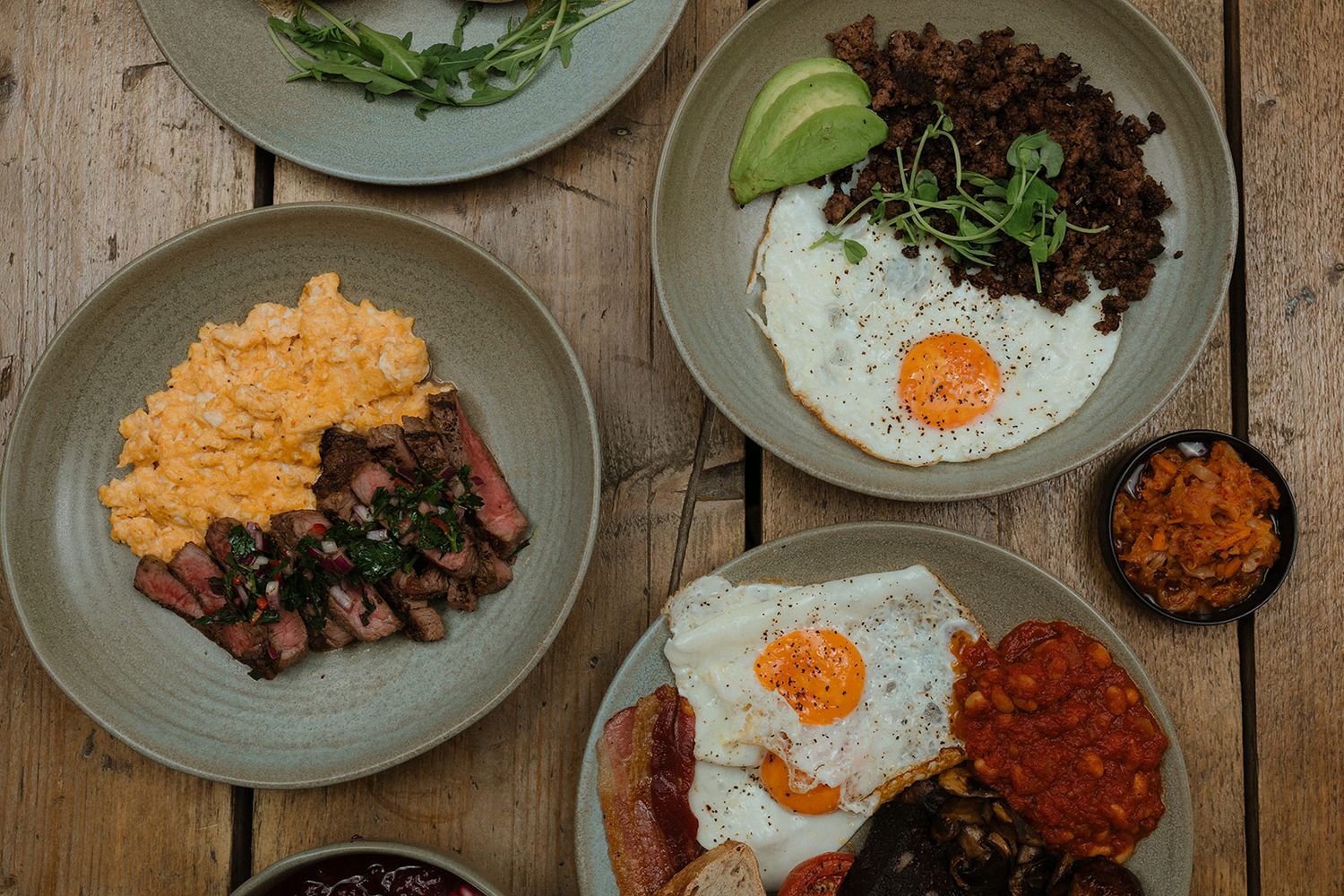 Overhead view of breakfast plates with fried eggs, steak, scrambled eggs, avocado, and sides on wooden table.