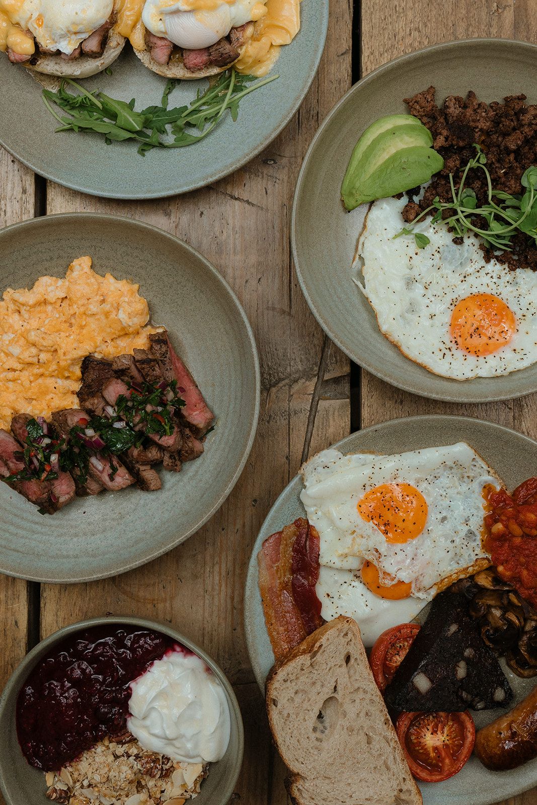 Overhead view of multiple breakfast plates featuring eggs, steak, avocado, and full English breakfast on wood table.