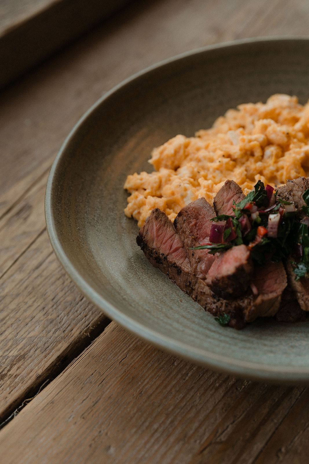 Tender sliced medium-rare steak with herb chimichurri and creamy scrambled eggs on a rustic ceramic plate.