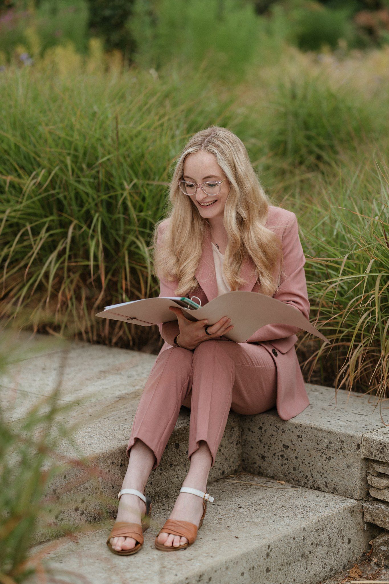 Smiling blonde woman in pink suit sitting on stone steps outdoors, reading a folder among lush green grasses.