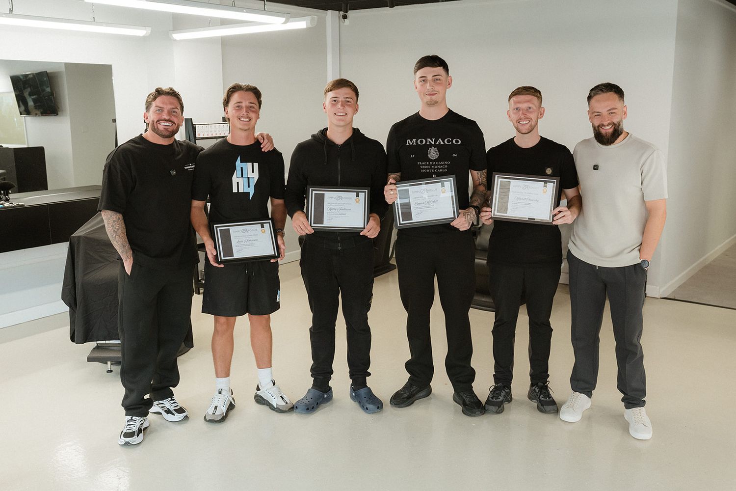 Six men in black attire proudly hold certificates while posing together in a modern barbershop setting.