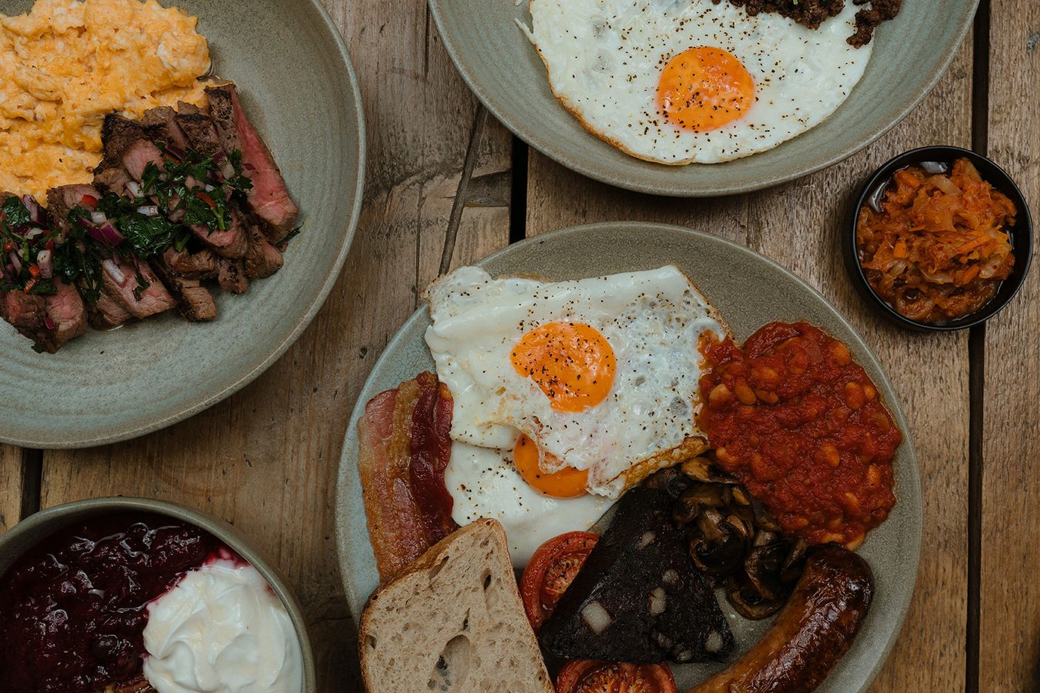 Overhead view of a full English breakfast with fried eggs, bacon, sausages, beans, and toast on wooden table.