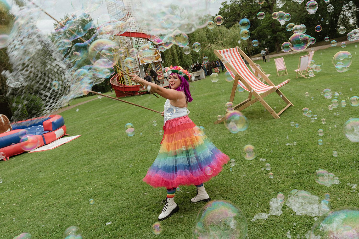 Woman in rainbow skirt playing with giant bubbles on a sunny lawn with deck chairs.