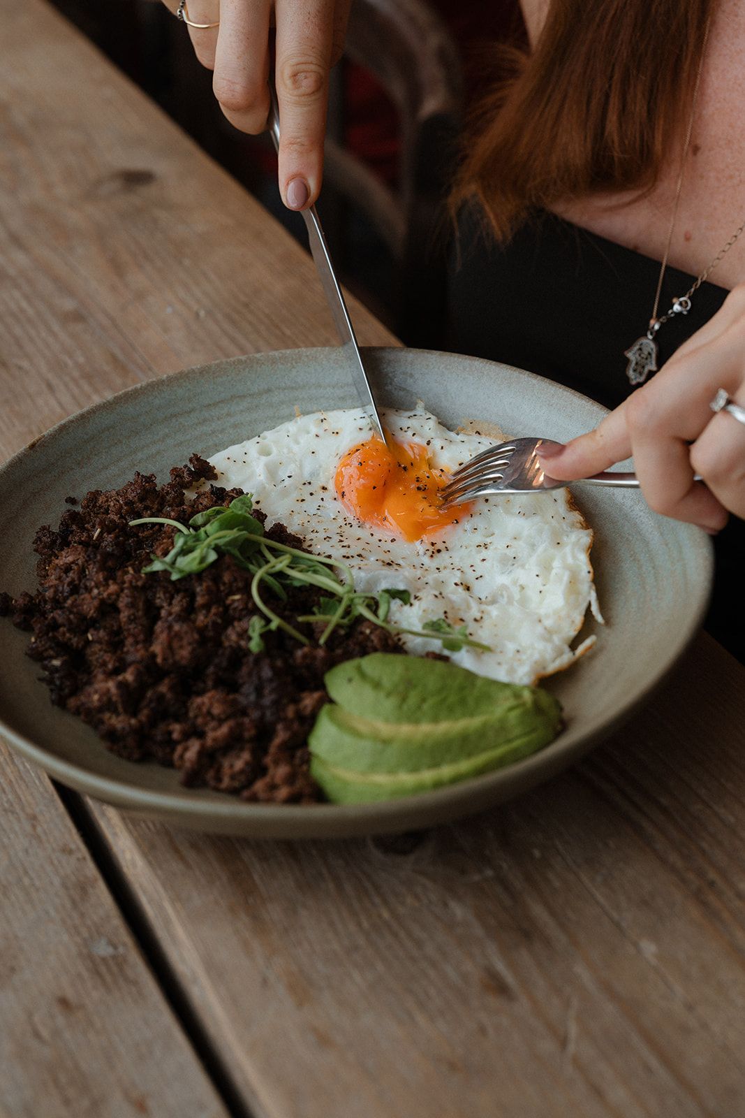 Woman cutting into a sunny-side-up egg bowl with black beans and avocado slices on a wooden table.