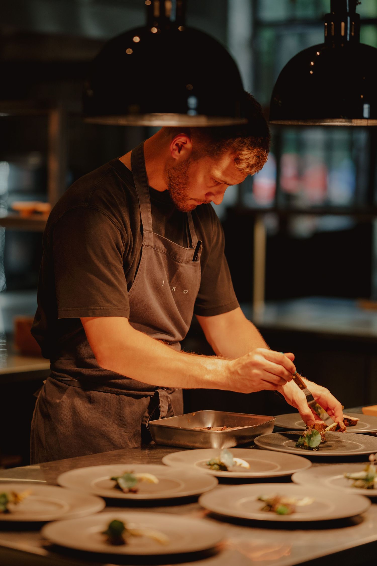 Chef carefully plates gourmet dishes under warm kitchen lights wearing a gray apron in a fine dining restaurant.