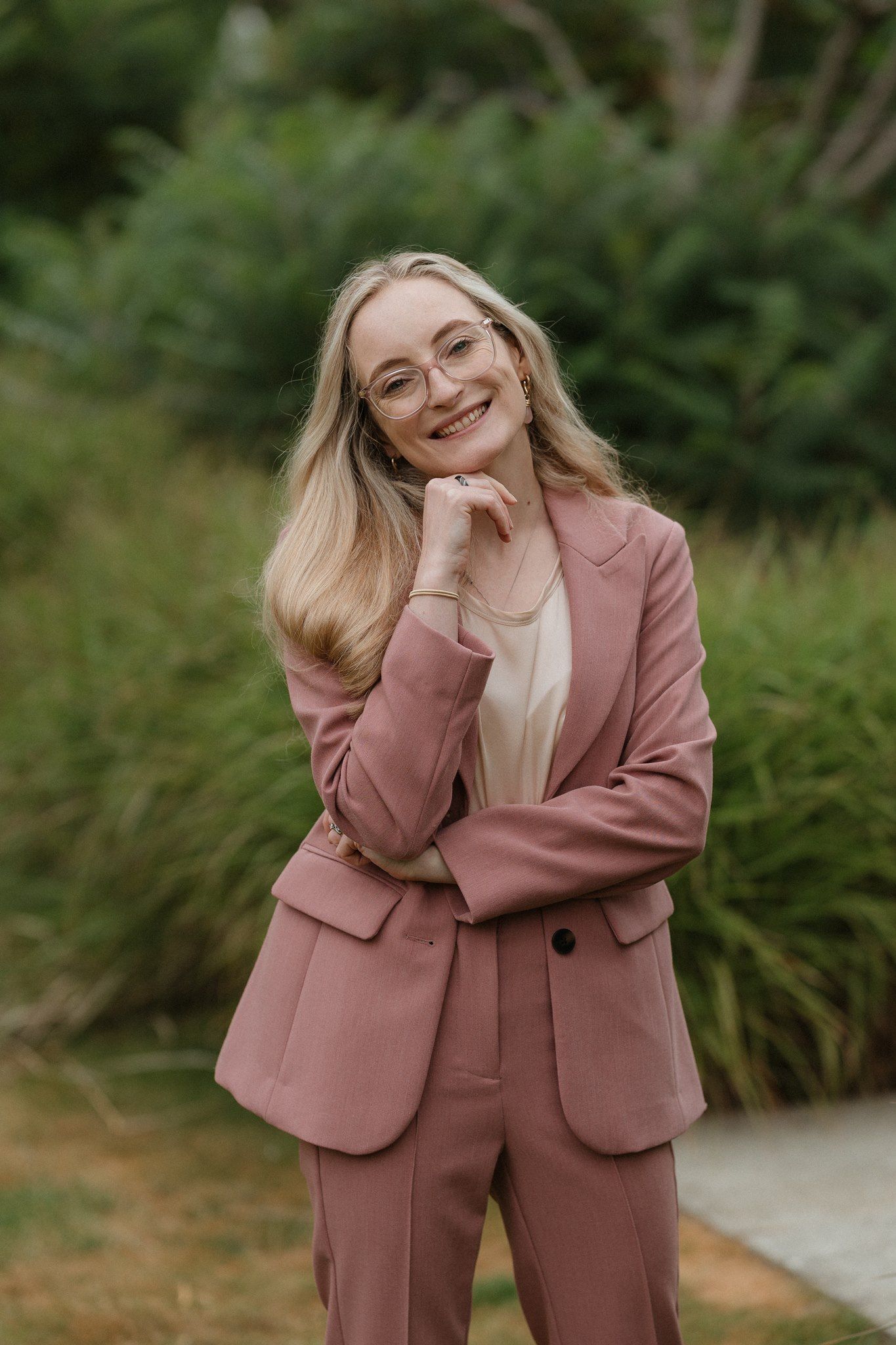 Smiling blonde woman in pink blazer and pants suit wearing glasses, posing outdoors with green shrubs background.