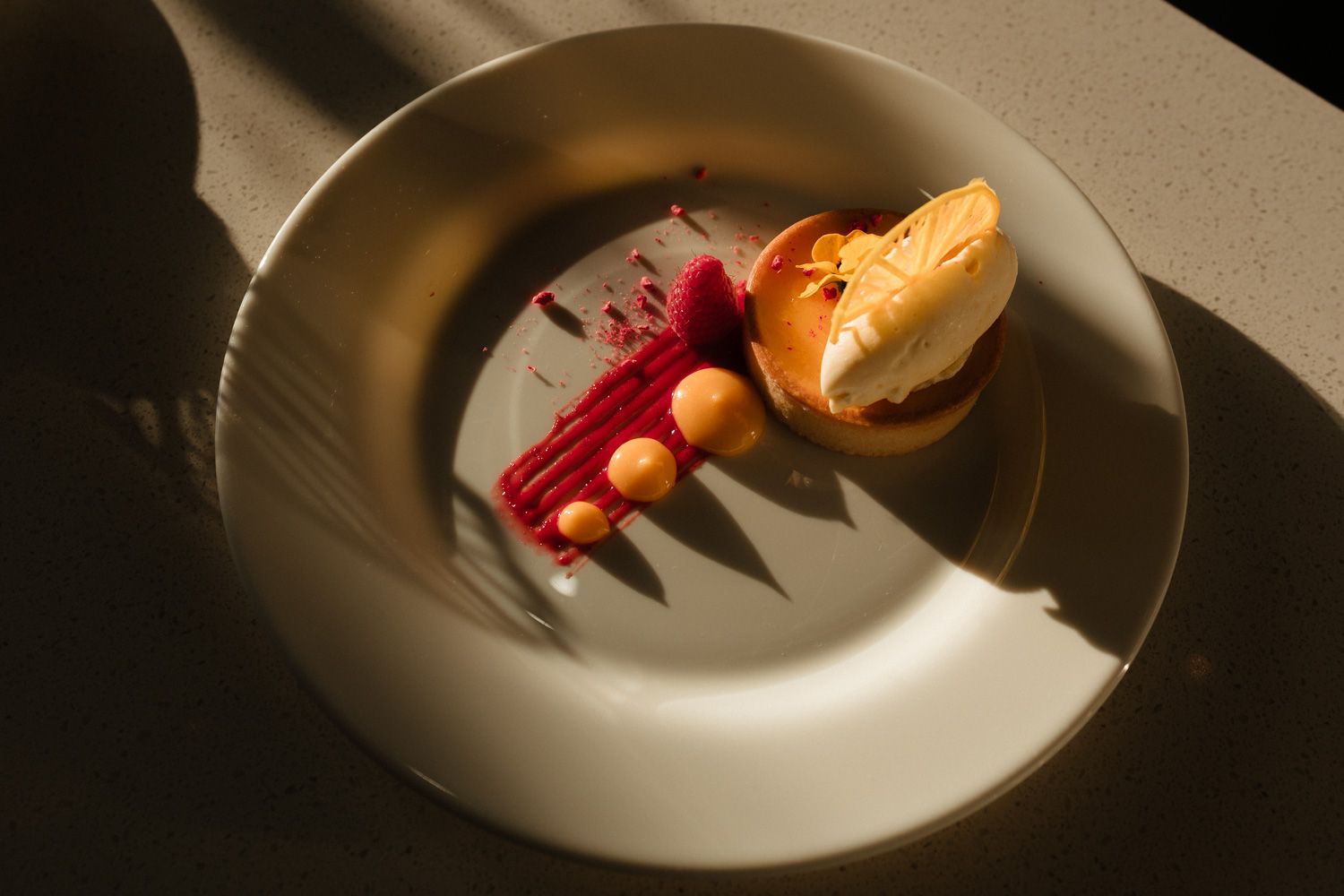 Elegant plated dessert with raspberry coulis, orange sorbet, and fruit pearls on a white bowl in dramatic light.