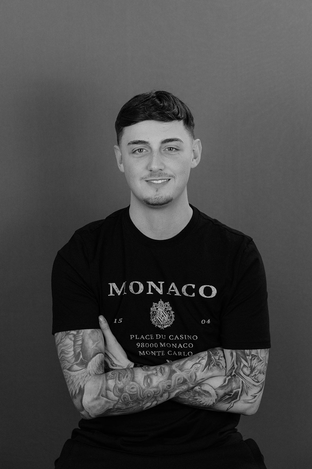 Black and white portrait of a young tattooed man with crossed arms wearing a Monaco t-shirt.