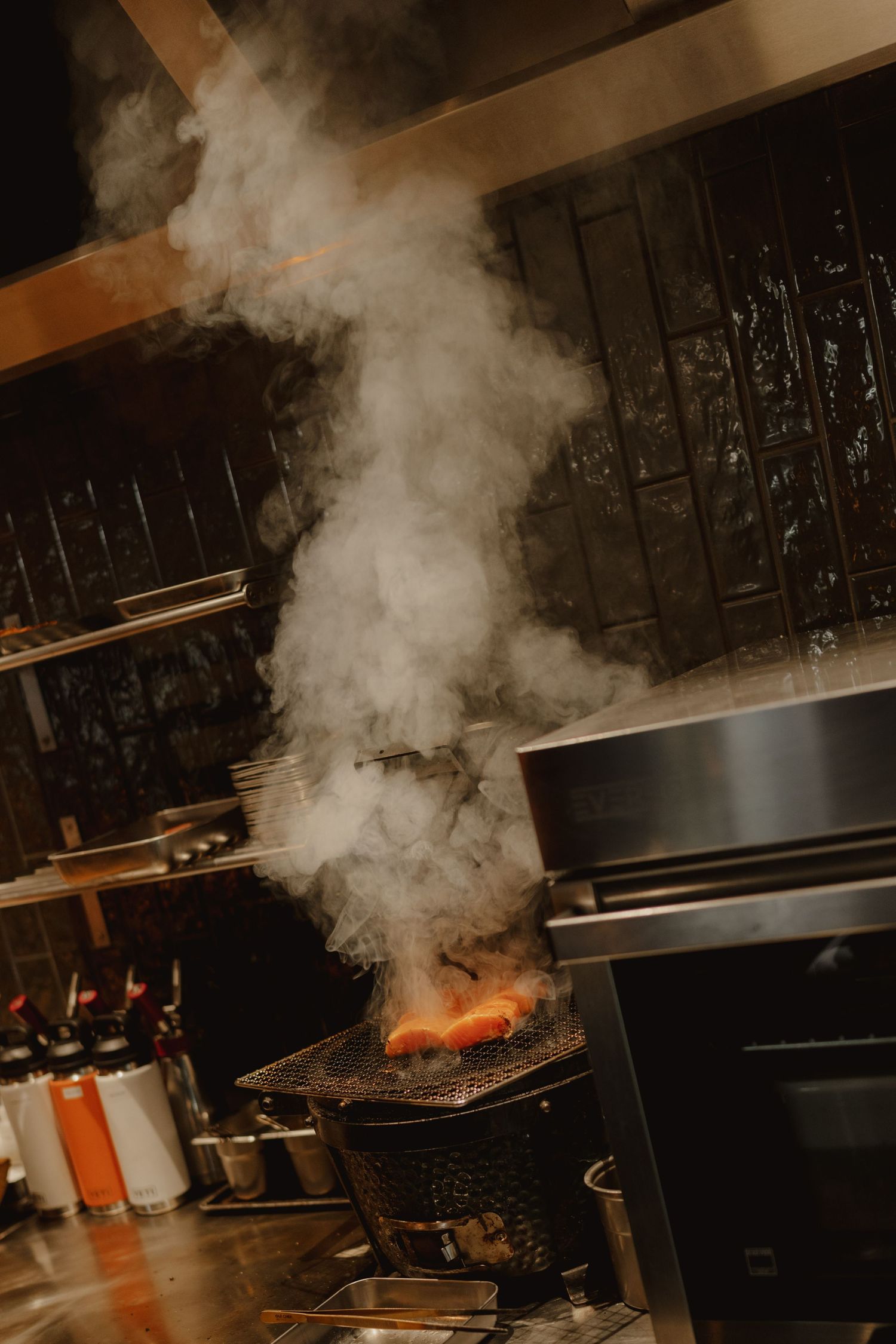 Smoke rising from flaming grill in a professional restaurant kitchen with stainless steel equipment.