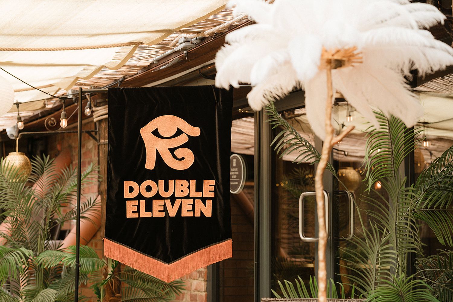 Double Eleven logo banner displayed outside a restaurant entrance surrounded by tropical plants and festive decorations.