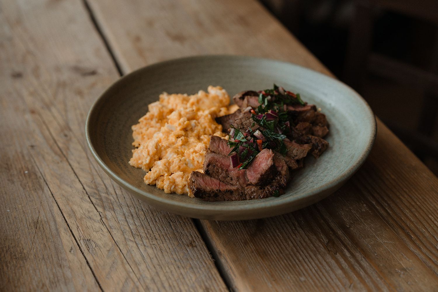 Sliced medium-rare steak with herb chimichurri alongside creamy scrambled eggs on a ceramic plate.