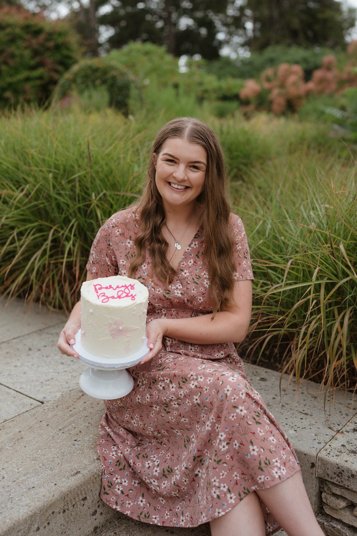 Young woman in floral dress holding white birthday cake outdoors in garden setting, smiling.