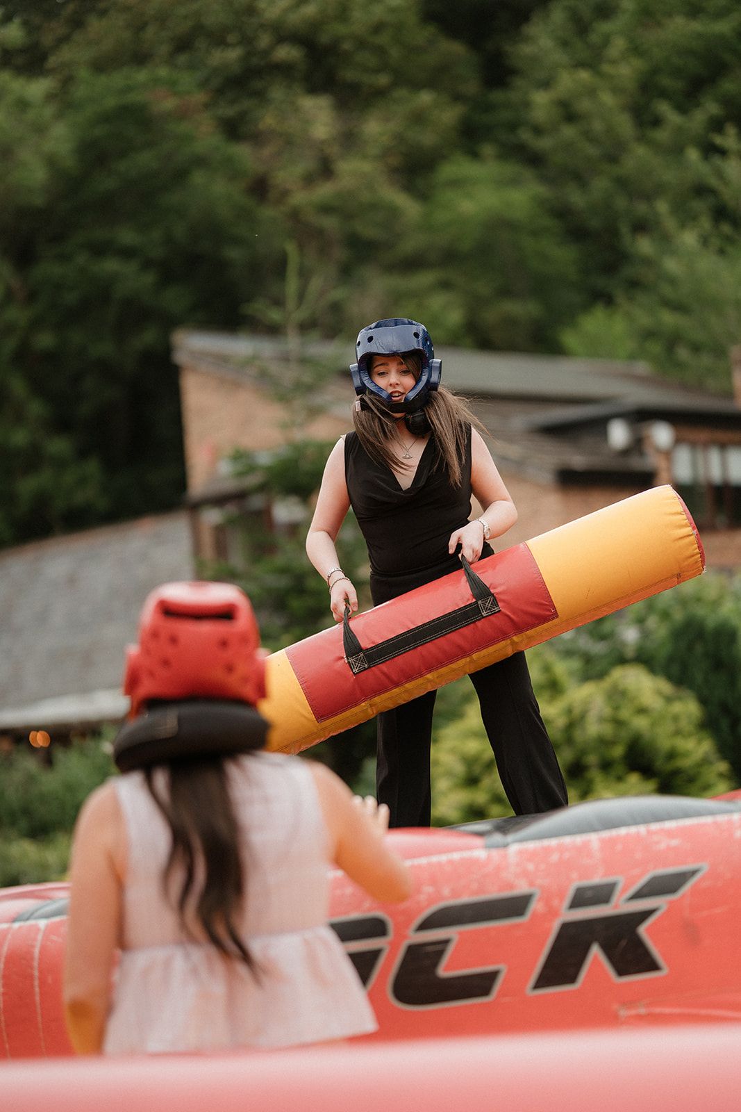Two women wearing helmets compete in an inflatable jousting game outdoors.
