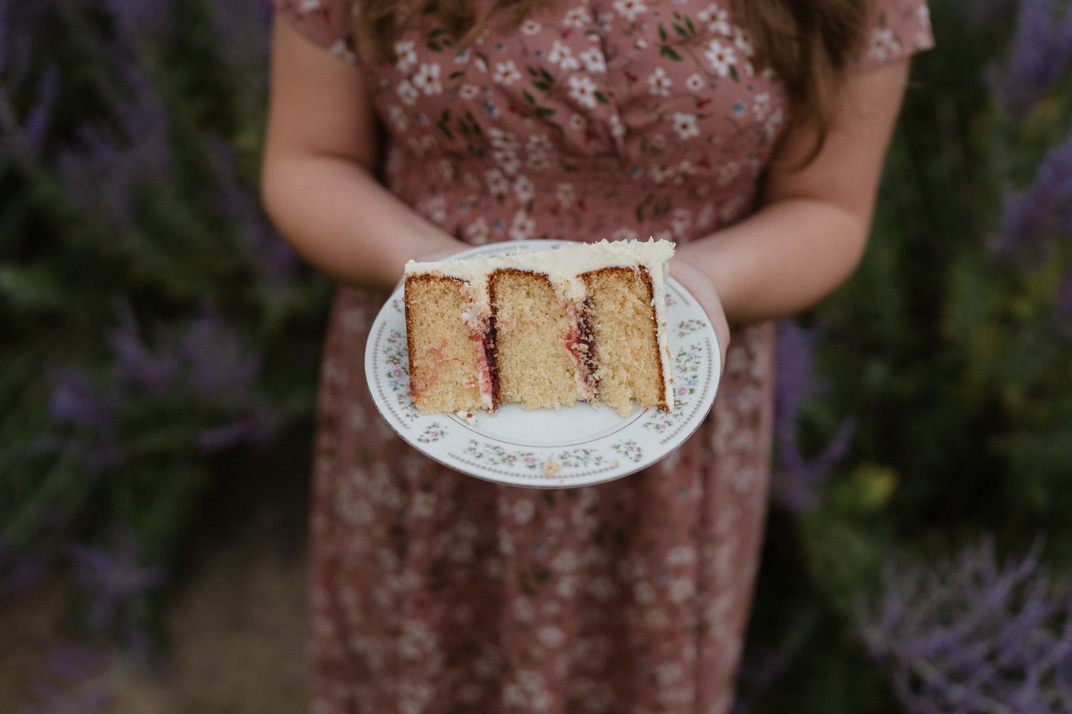 Woman in floral dress holding a white plate with sliced sponge cake with jam filling outdoors.