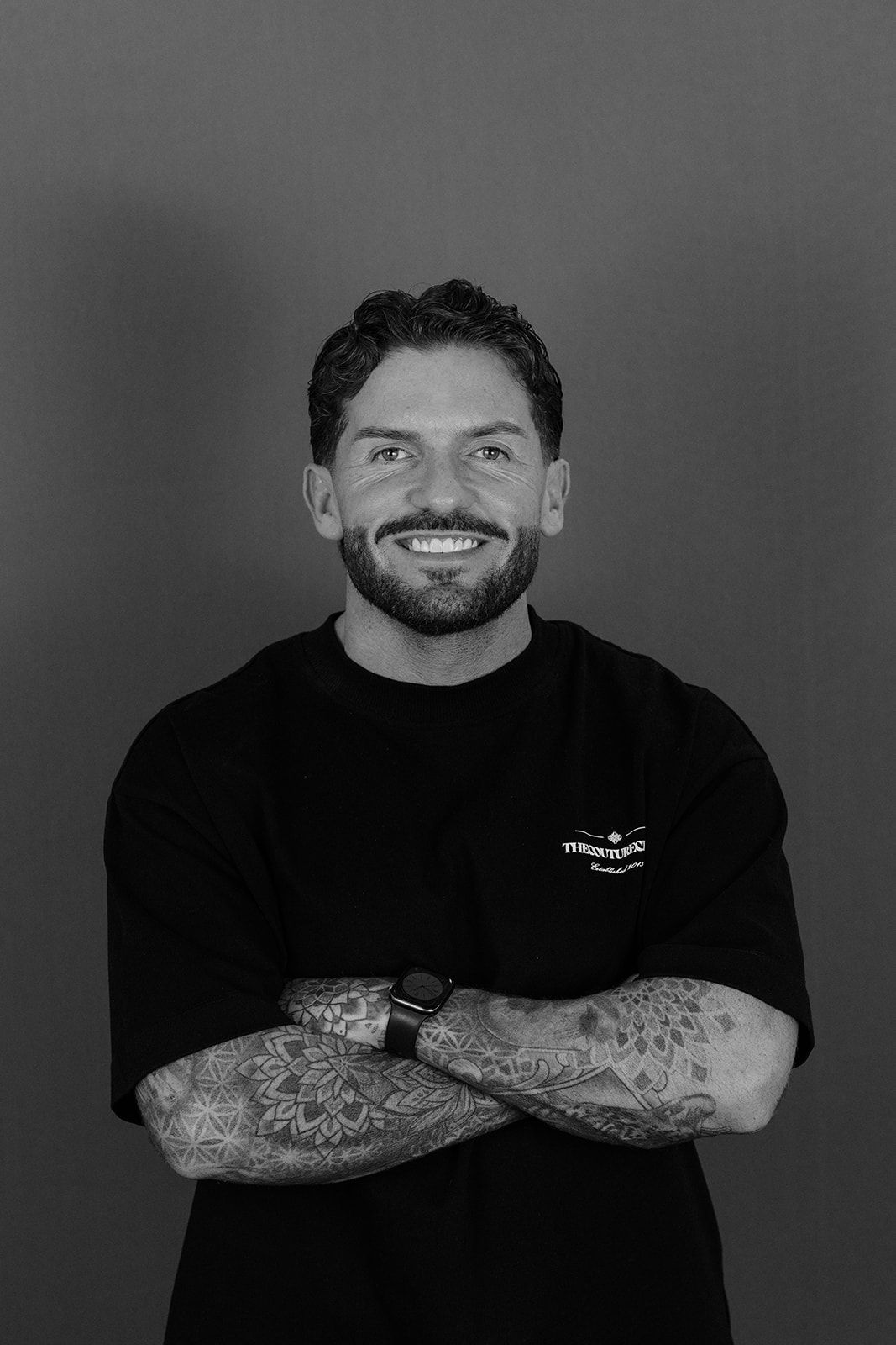 Smiling bearded man with tattoos crosses arms wearing black t-shirt in black and white studio portrait.