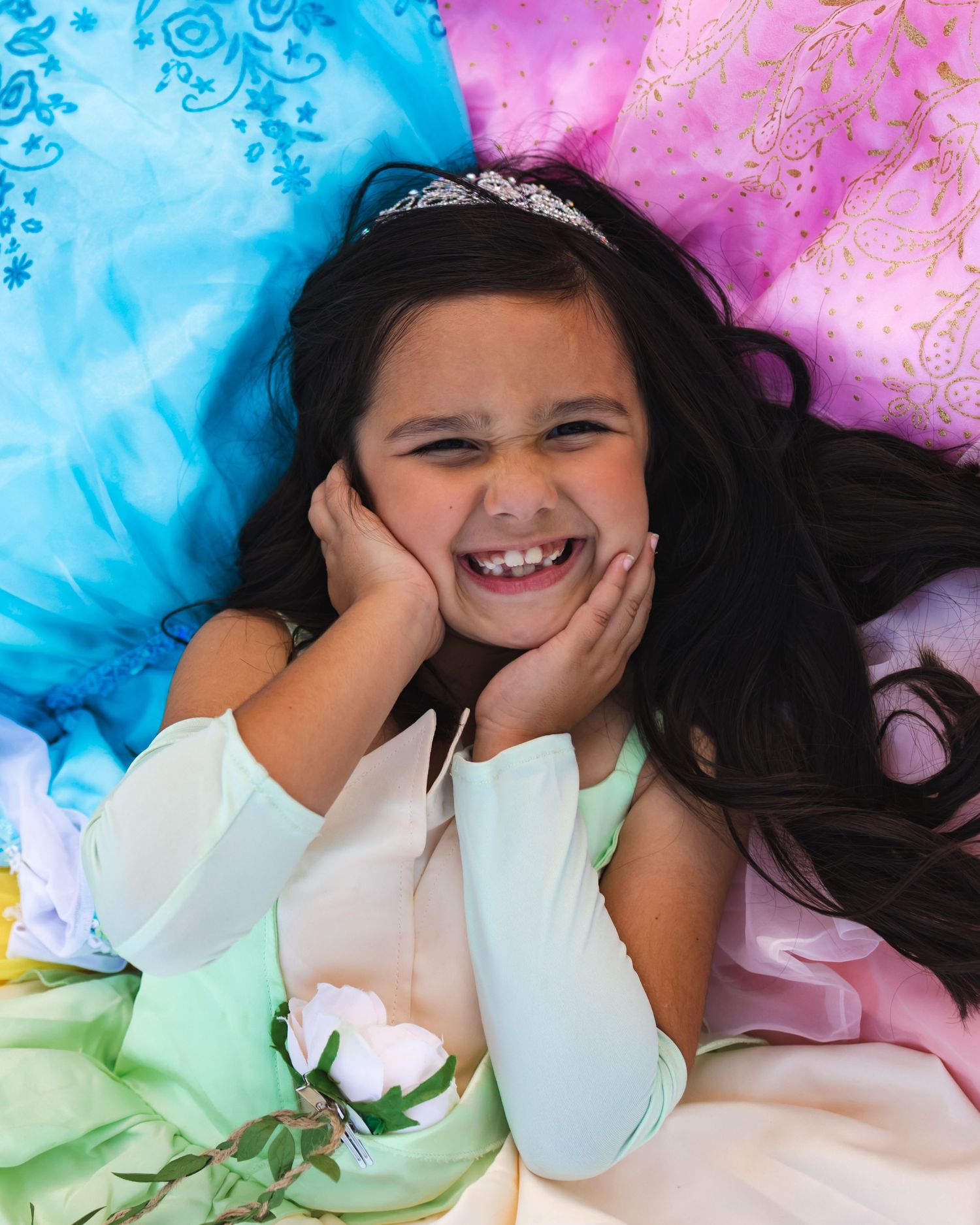 A joyful young girl in a mint green dress poses with hands on cheeks, surrounded by colorful princess gowns.