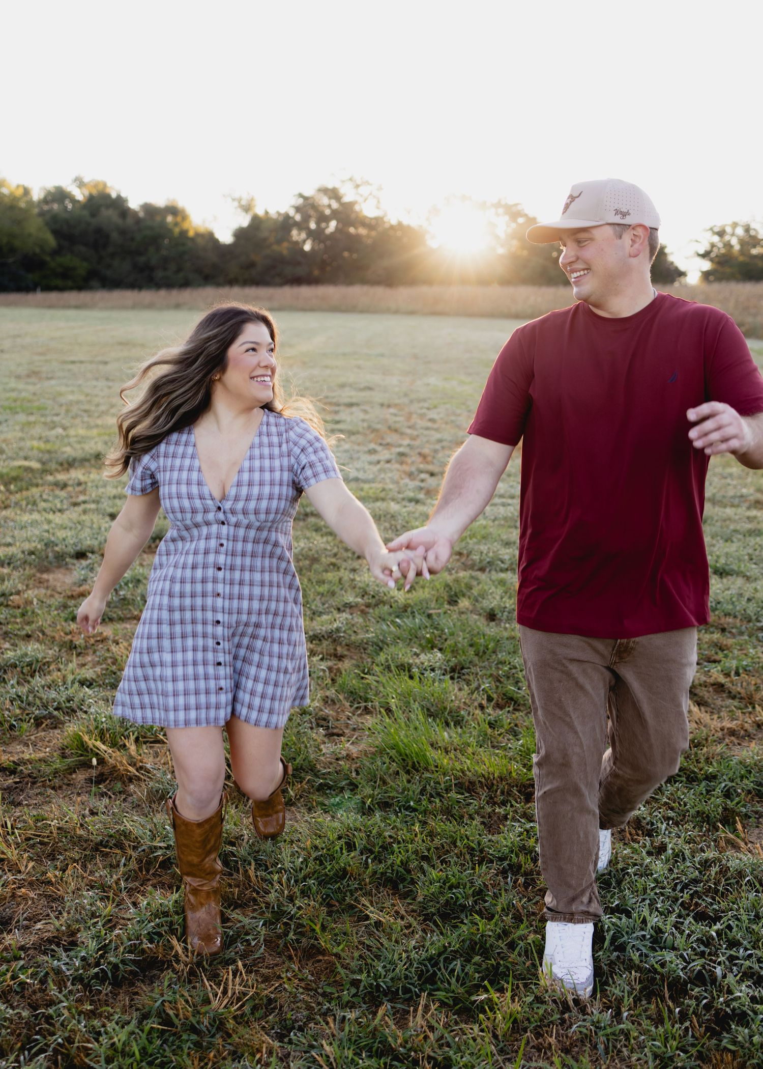 A smiling couple holds hands while walking through a sunlit field at golden hour, she in a plaid dress and boots.