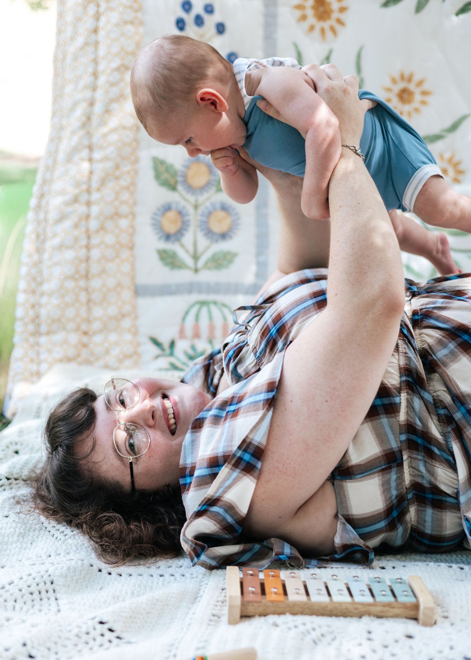 Parent lying down lifting smiling baby in blue onesie while playing on a floral quilt indoors.