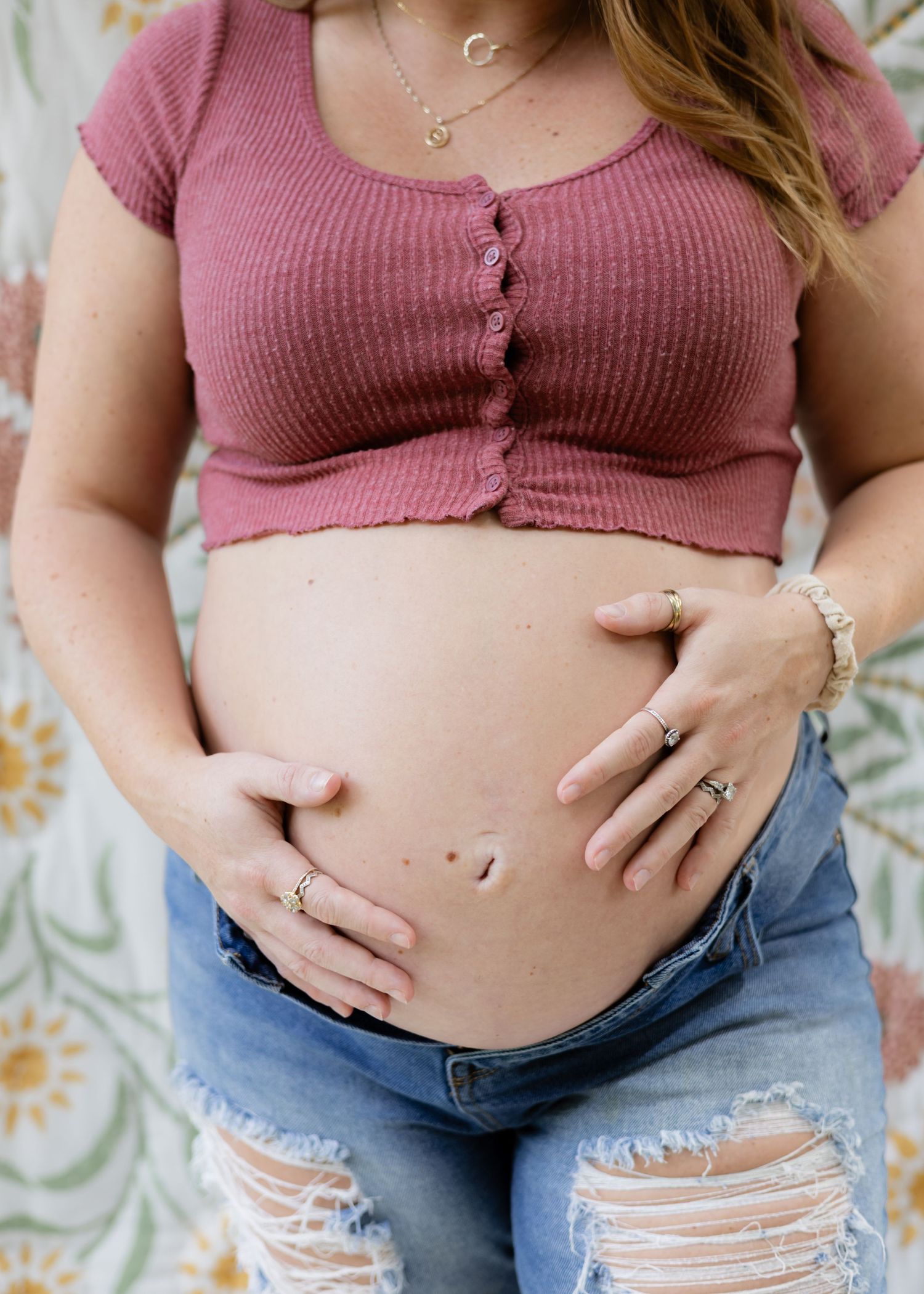 Pregnant woman in pink crop top and ripped jeans cradling her bare baby bump with both hands.