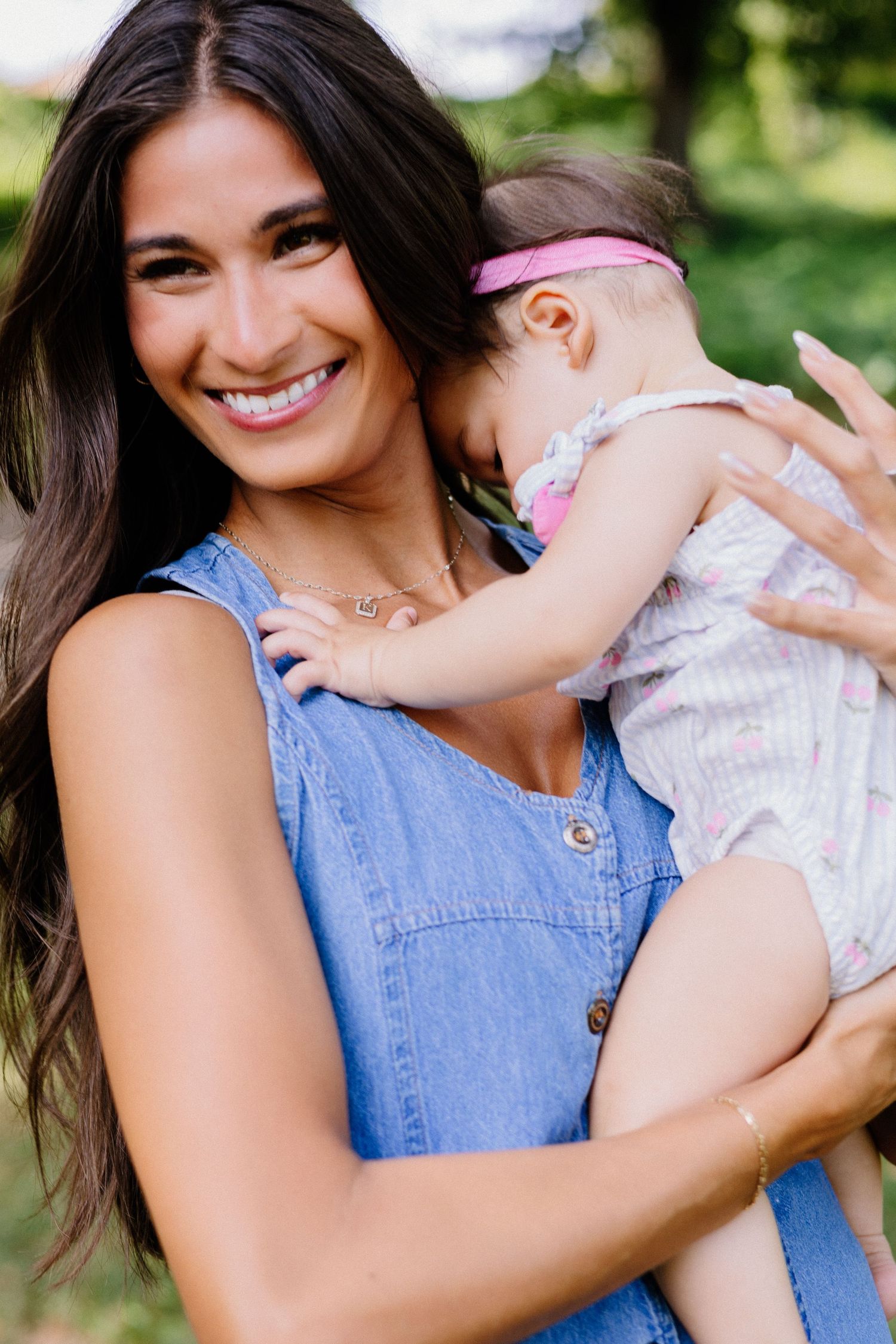 Smiling brunette woman in denim dress holding a baby girl wearing a pink headband outdoors.