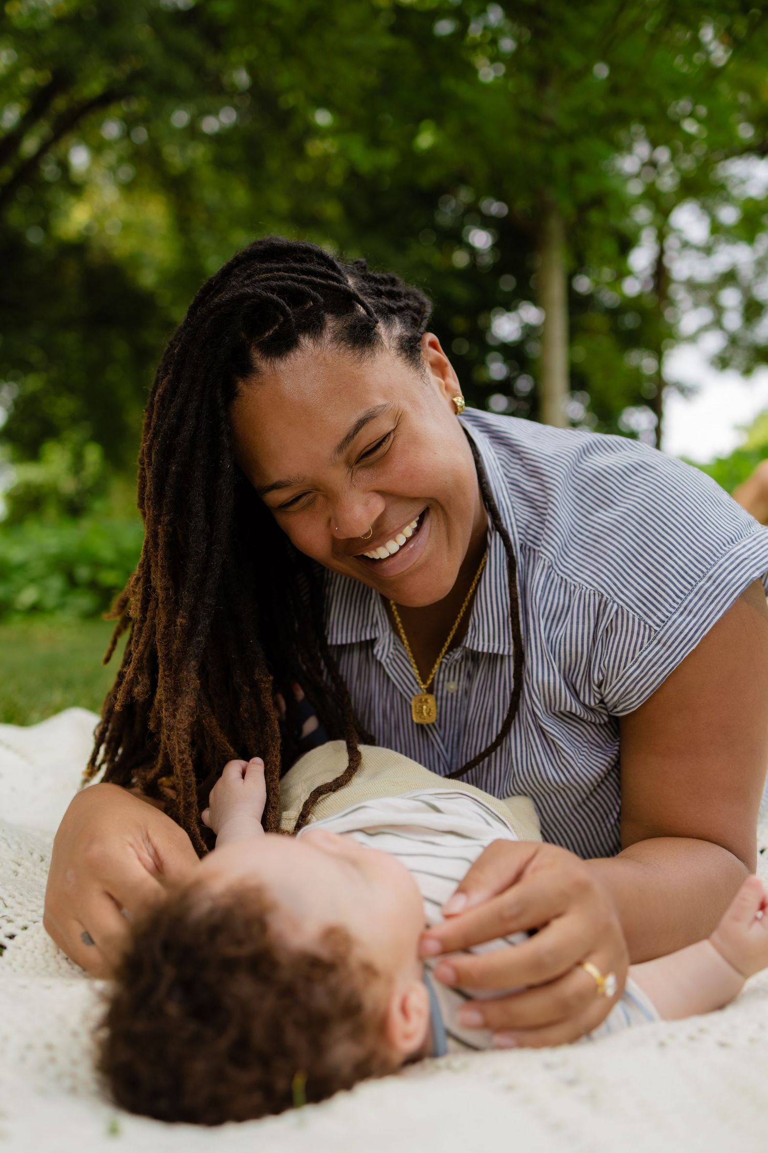 Laughing mother with dreadlocks lying on blanket outdoors smiling joyfully at her baby.