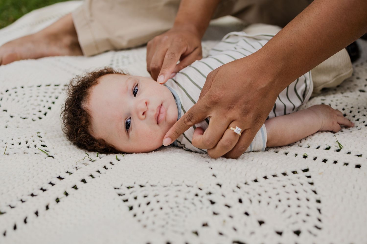 Newborn baby in striped onesie lying on white blanket while mother's hands gently hold them.