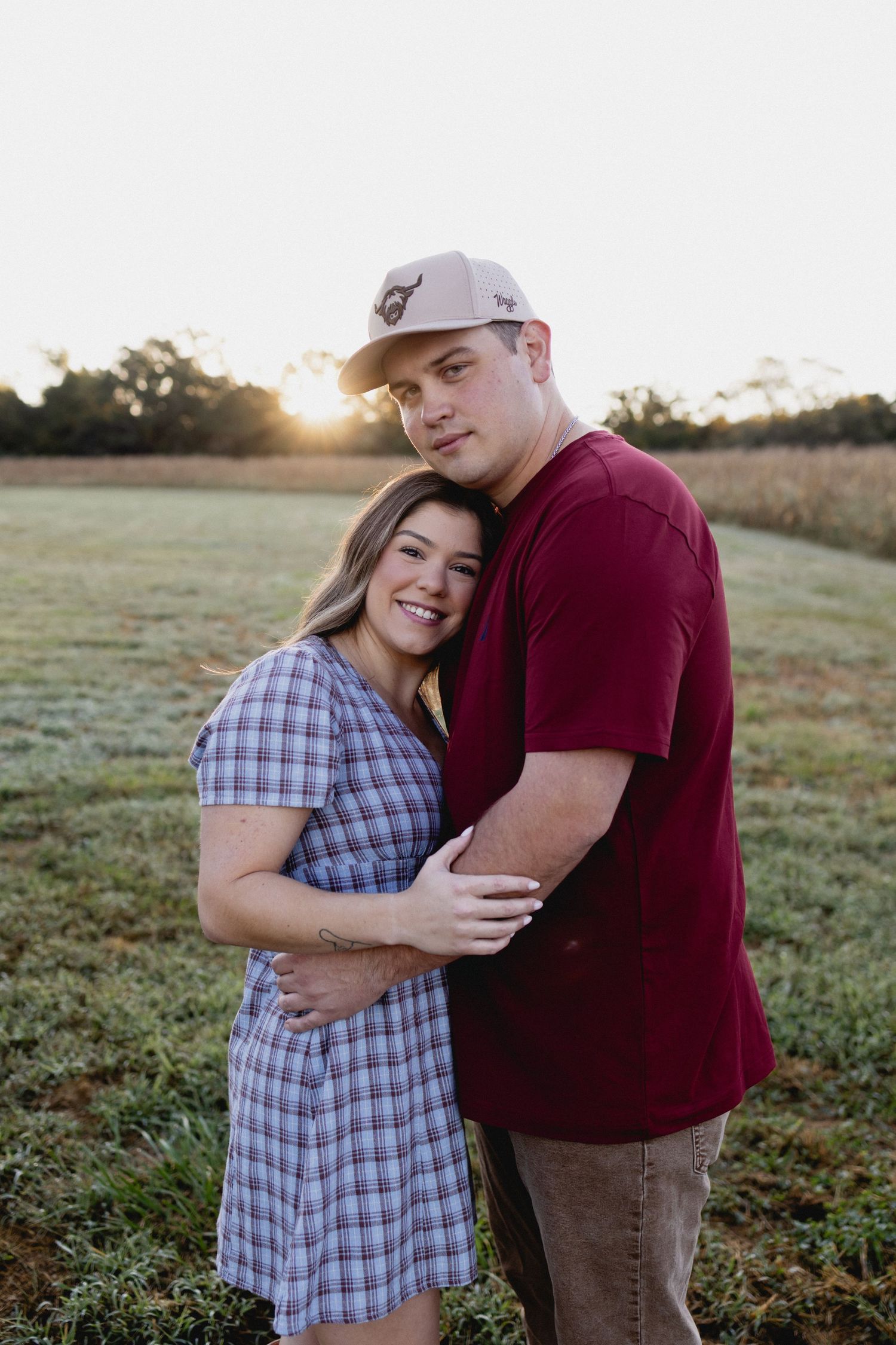 A smiling couple embraces in a golden field at sunset, woman in plaid dress, man in maroon shirt and cap.