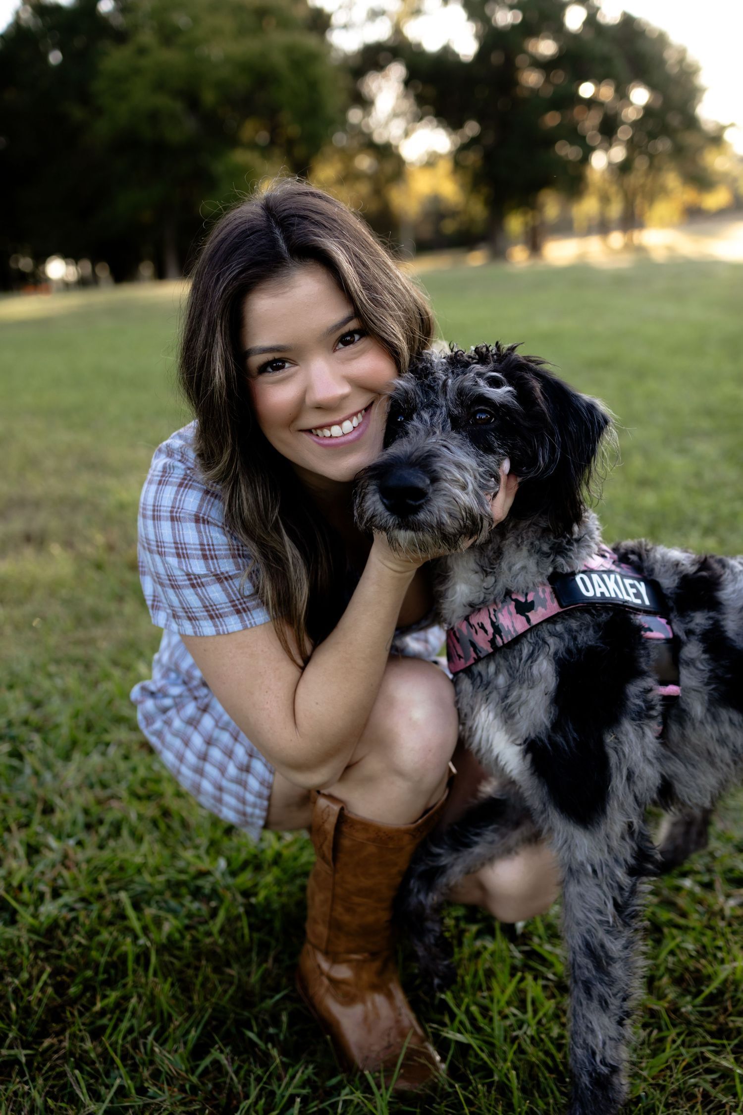 Young woman in plaid shirt and cowboy boots smiles while hugging her black and gray dog named Oakley outdoors.