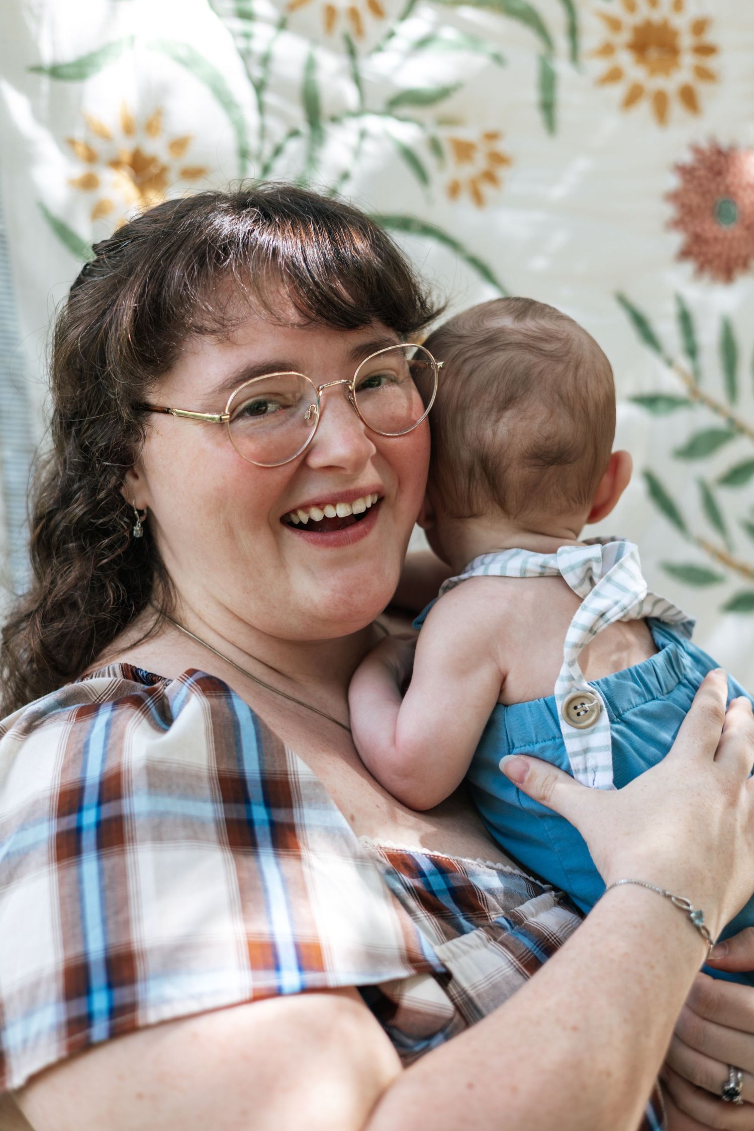 Happy woman with glasses in plaid shirt holding a newborn baby against a floral backdrop.