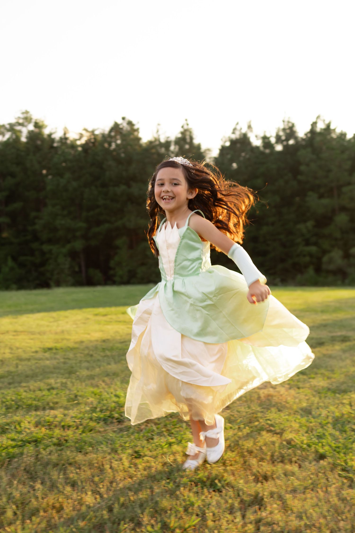 Young girl in green and yellow princess dress running joyfully on grass field with trees in background.