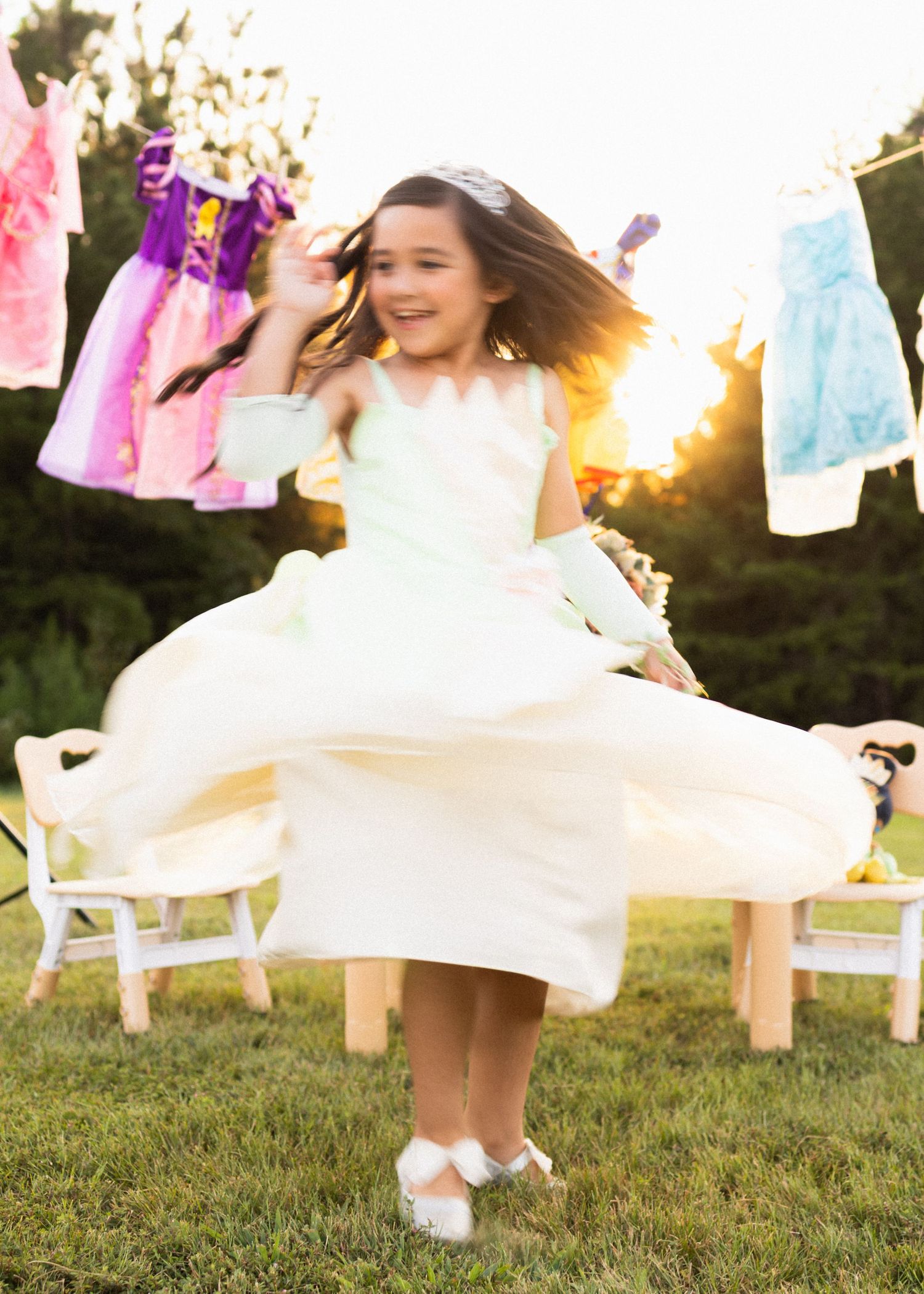A young girl twirls in a white dress outdoors at sunset, with colorful dresses hanging on a clothesline behind her.