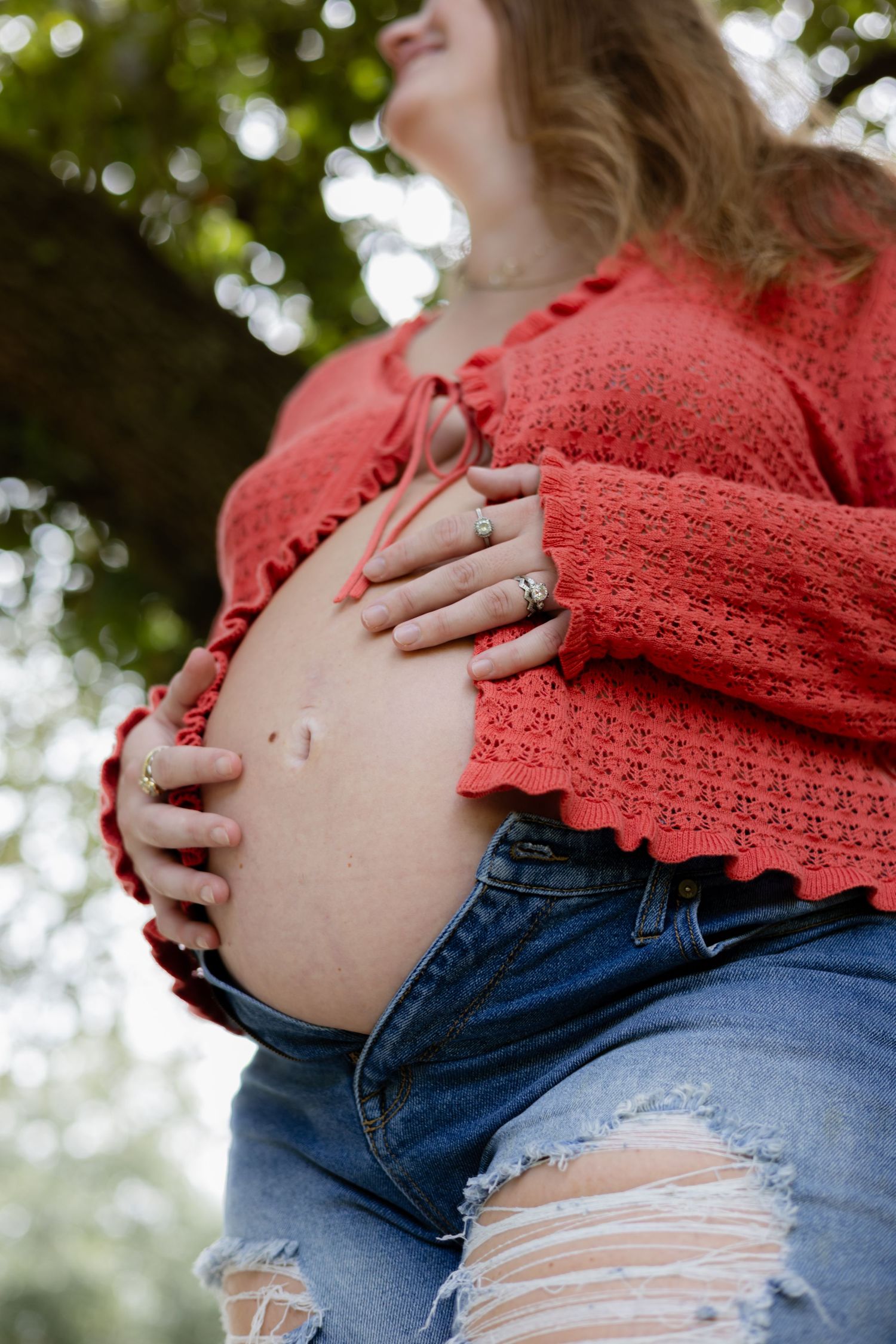 Close-up of pregnant woman in red lace top and distressed jeans holding her bare baby bump outdoors.