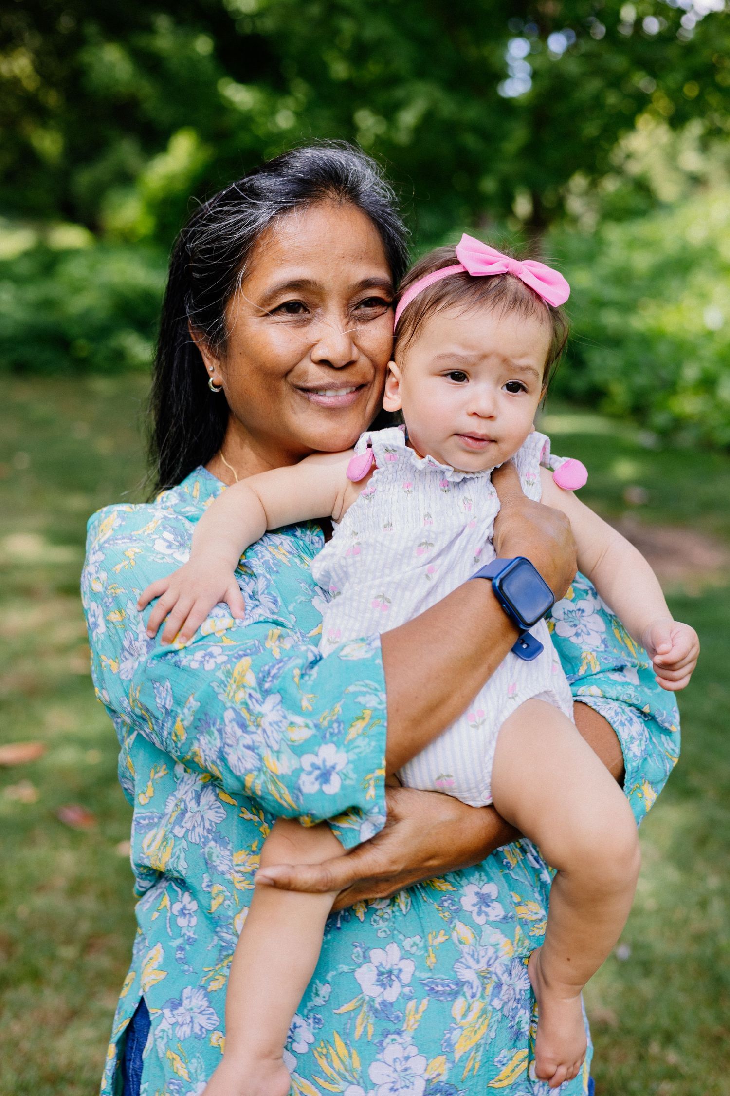 Smiling older woman in floral dress holding a baby girl with pink bow in a park.