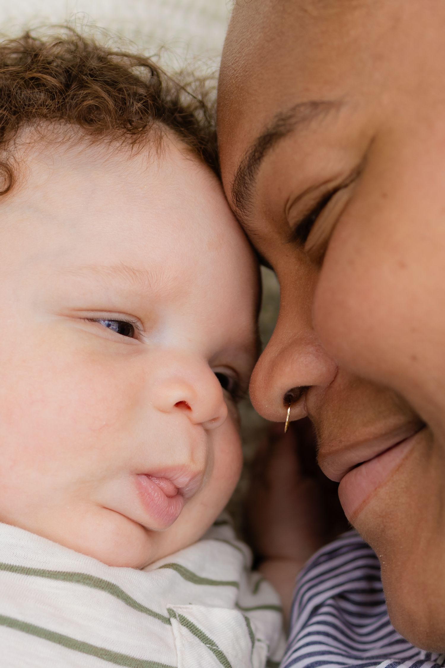 Close-up of smiling mother with nose ring cuddling her curly-haired newborn baby cheek to cheek.