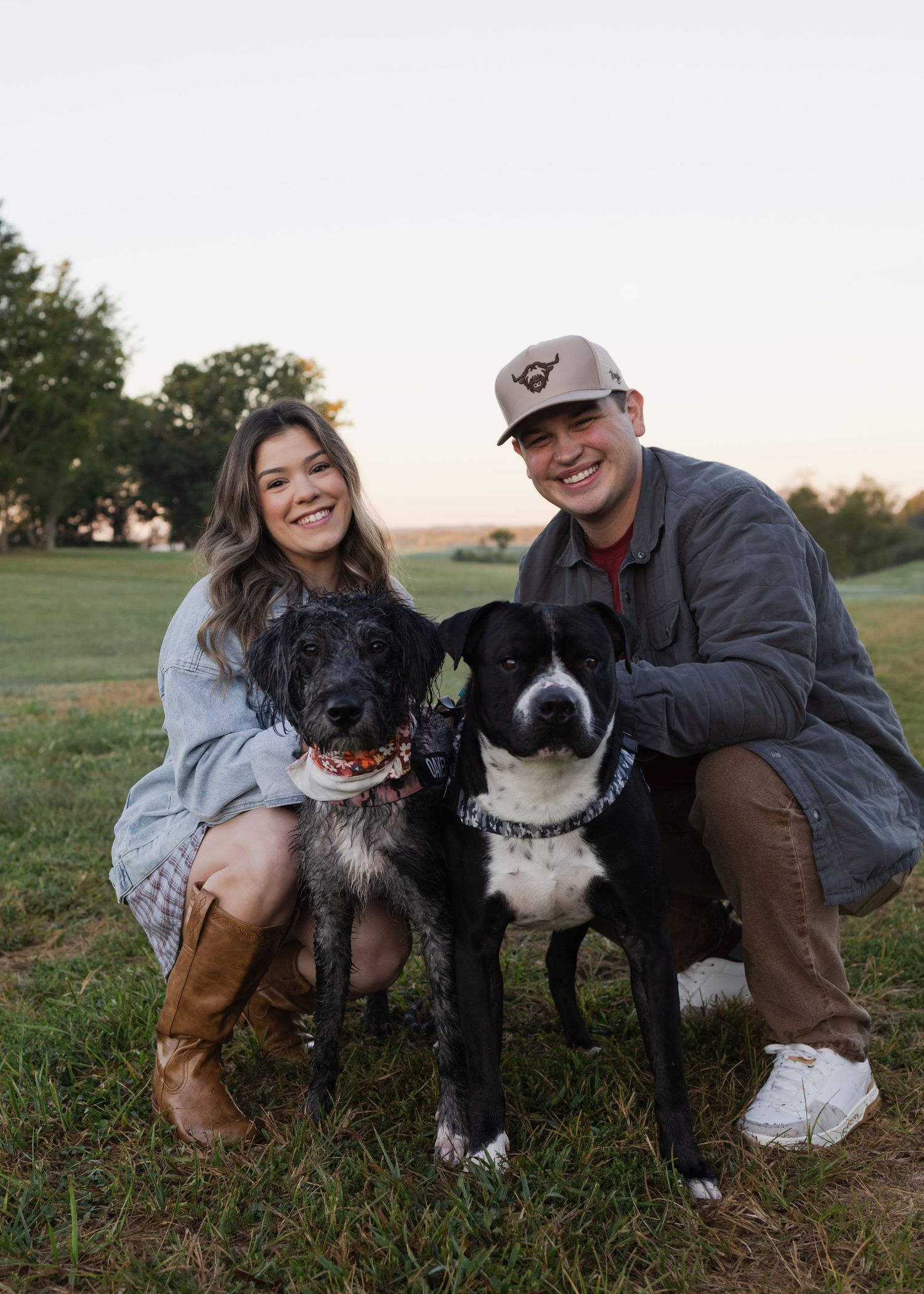 A smiling couple crouches in a grassy field at dusk with two dogs, a brindle and a black-and-white, between them.