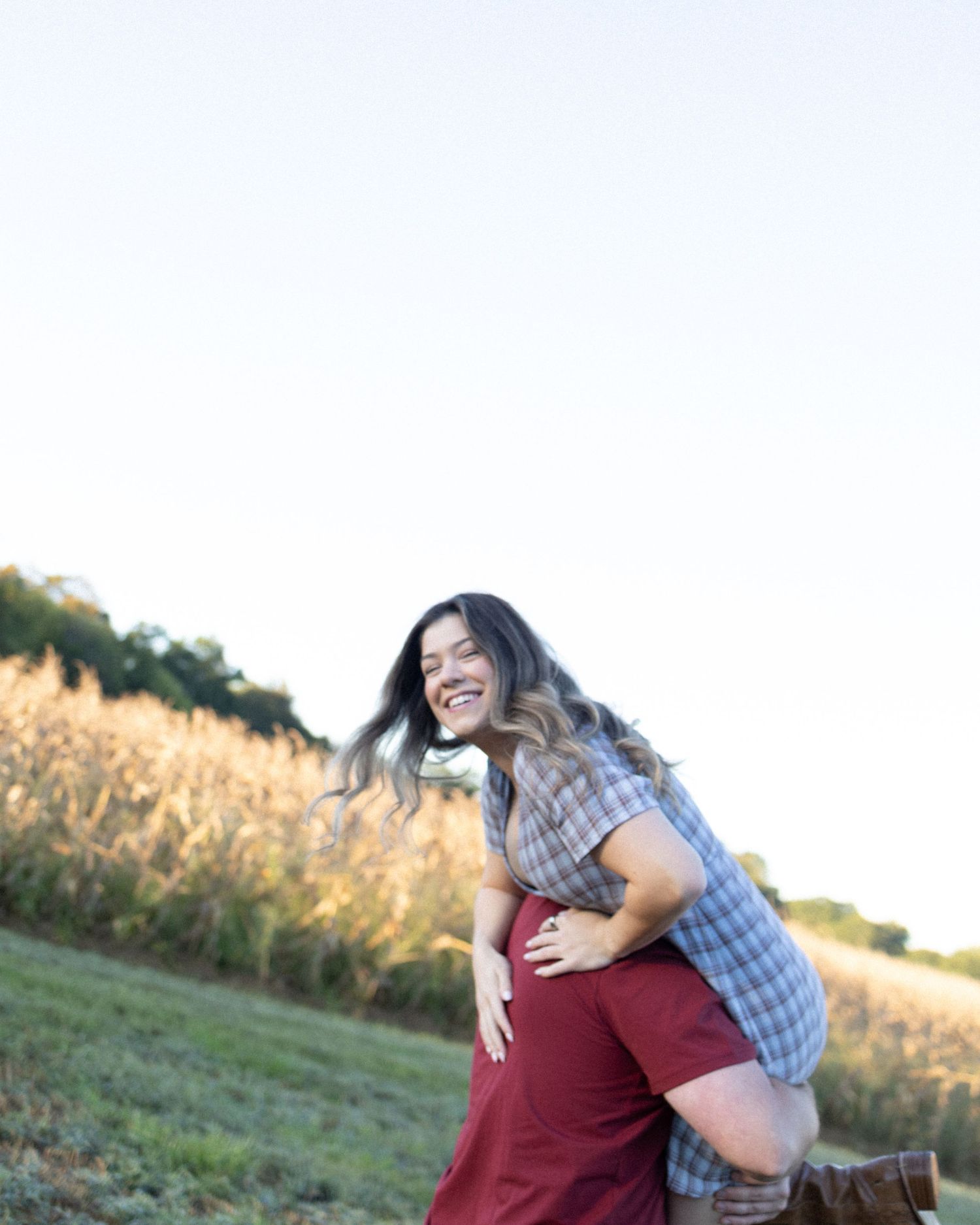 A laughing woman with long hair is joyfully lifted by a partner in a sunny field with golden crops.