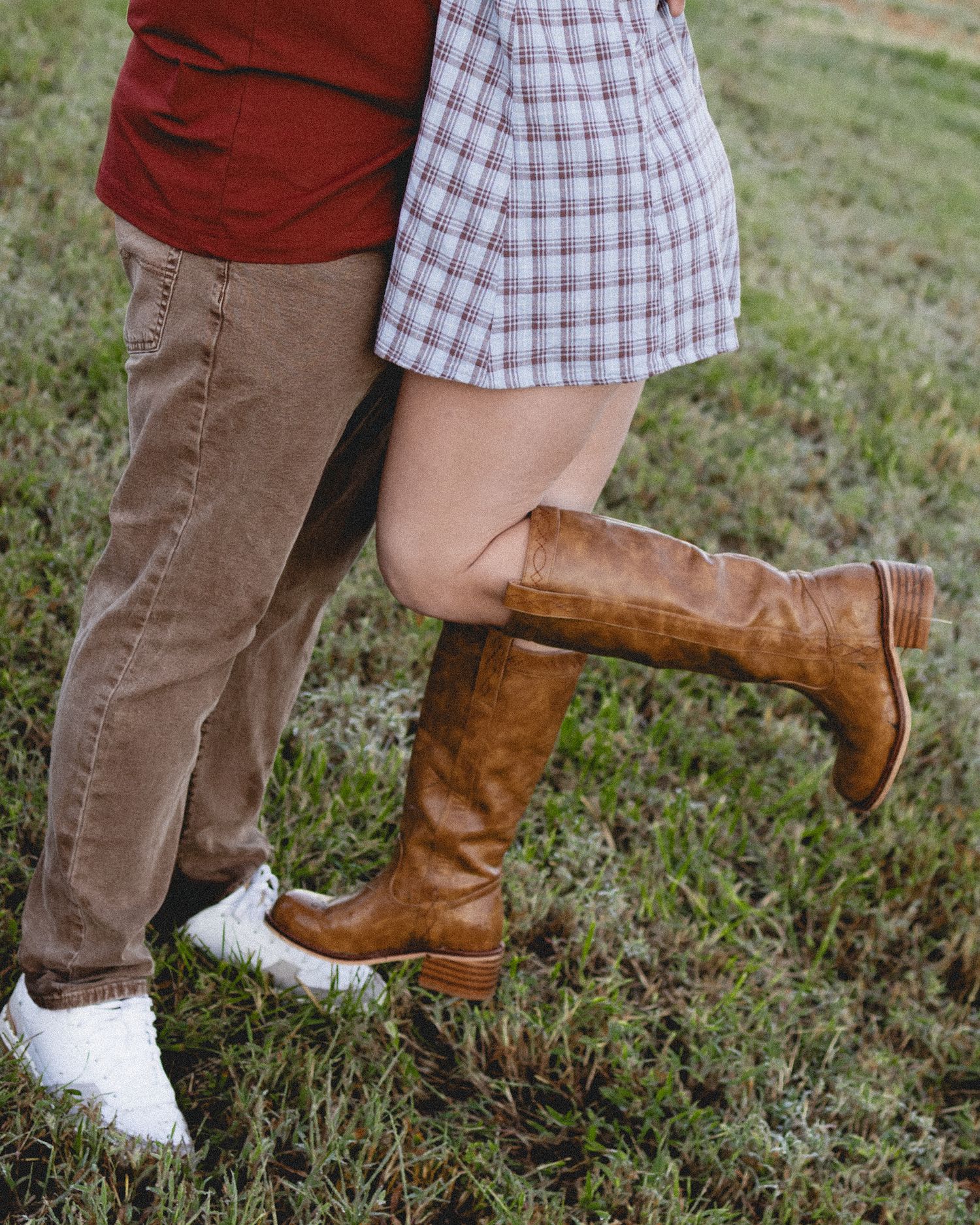 Couple standing close together on grass, woman wearing brown knee-high boots and plaid skirt, man in khakis and white sneakers.