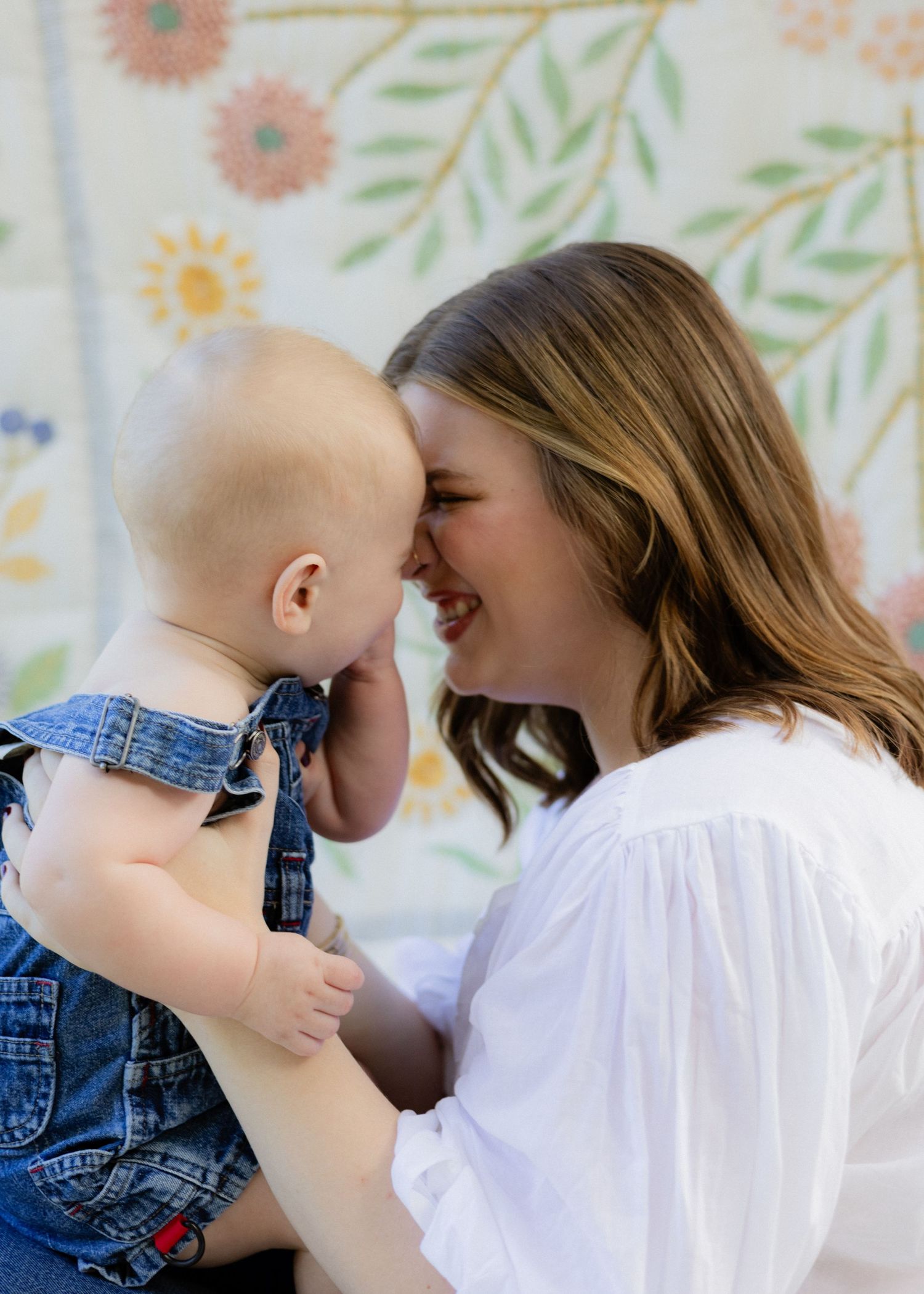 Happy mother in white dress touching foreheads with her baby in denim overalls near floral wall.