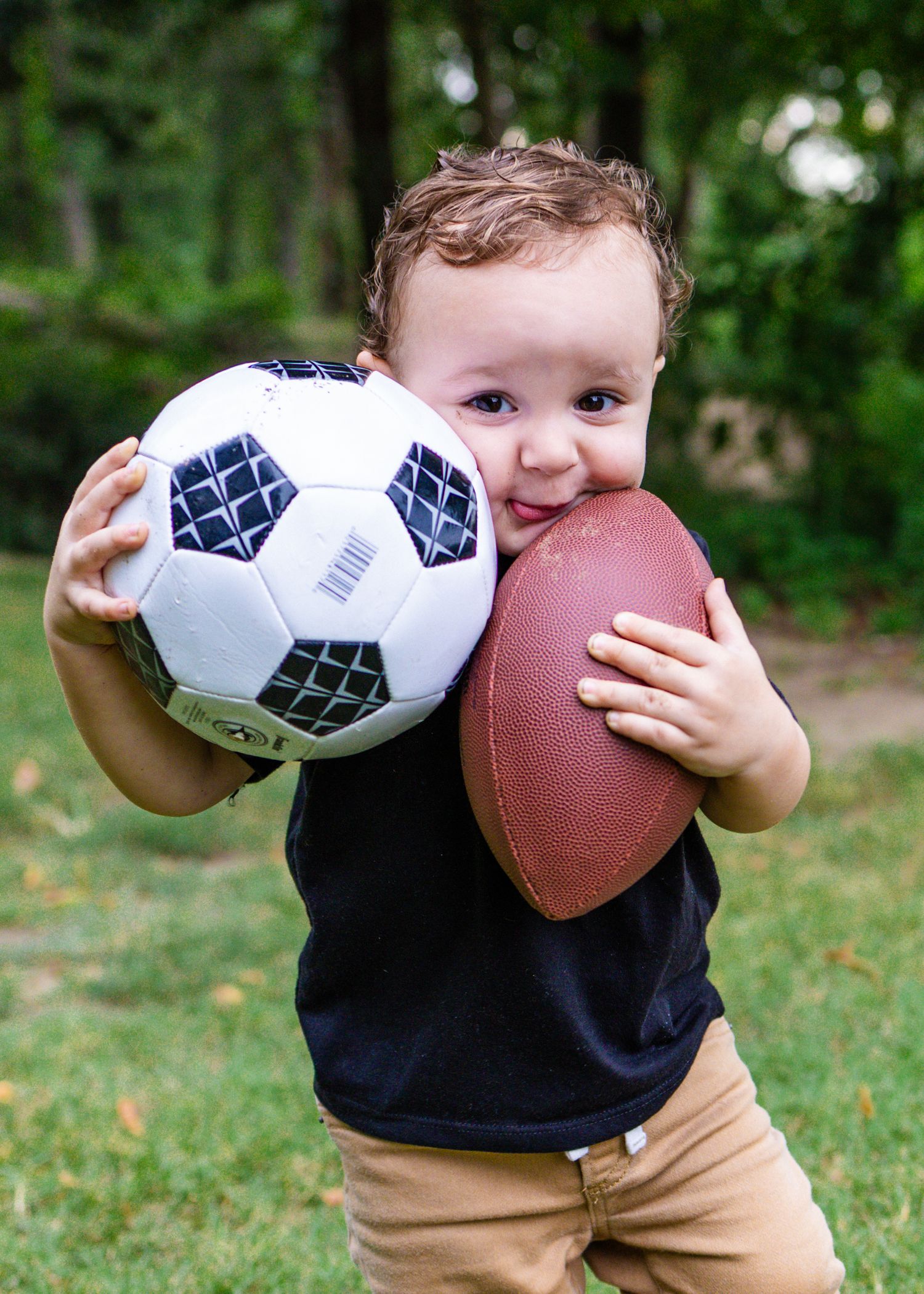 Adorable toddler boy holding a soccer ball and football while smiling outdoors in a park.