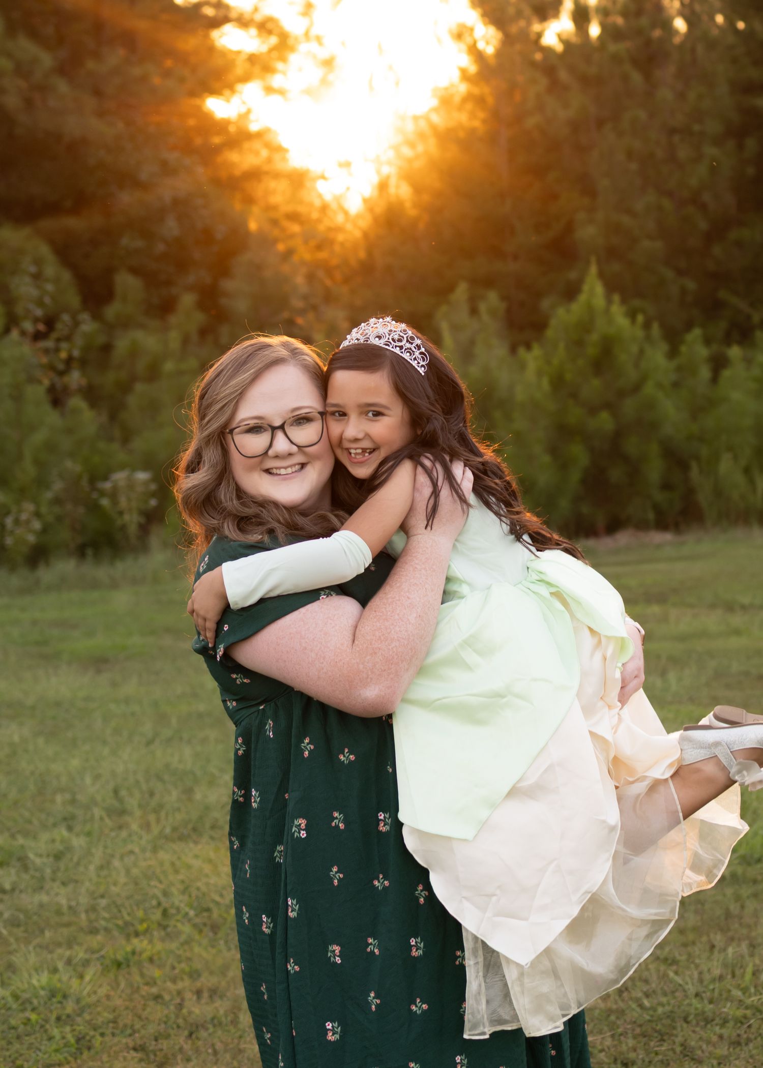 A teenage girl holds a young girl in a princess dress and tiara during a golden sunset outdoor photo session.